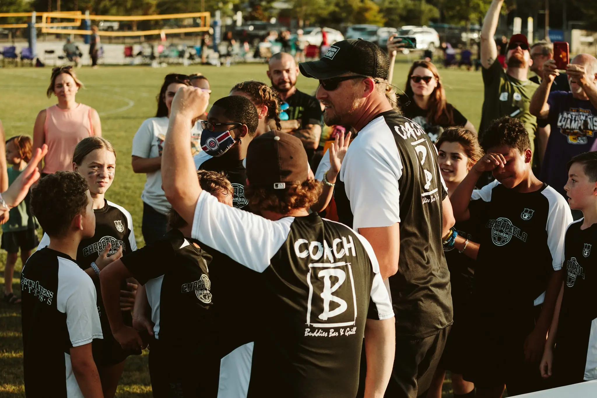 Youth Chappapeela Sports Park soccer players in black and white uniforms gather around two coaches on a grassy field during sunset.