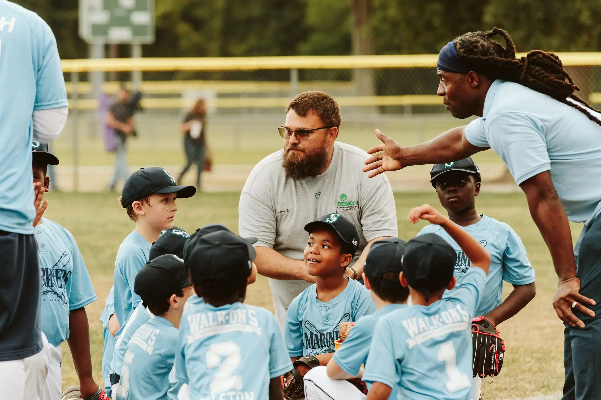 A H.A.R.D. #1 youth baseball coach with long dreadlocks and a headband giving instructions to a group of children in blue uniforms during practice.