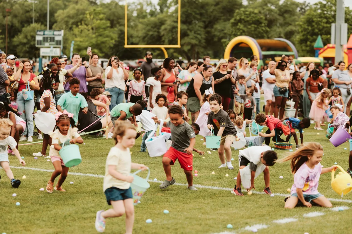 Children eagerly gathering Easter eggs in baskets on a grassy field during H.A.R.D. #1's Easter Eggstravaganza with a crowd of spectators watching.