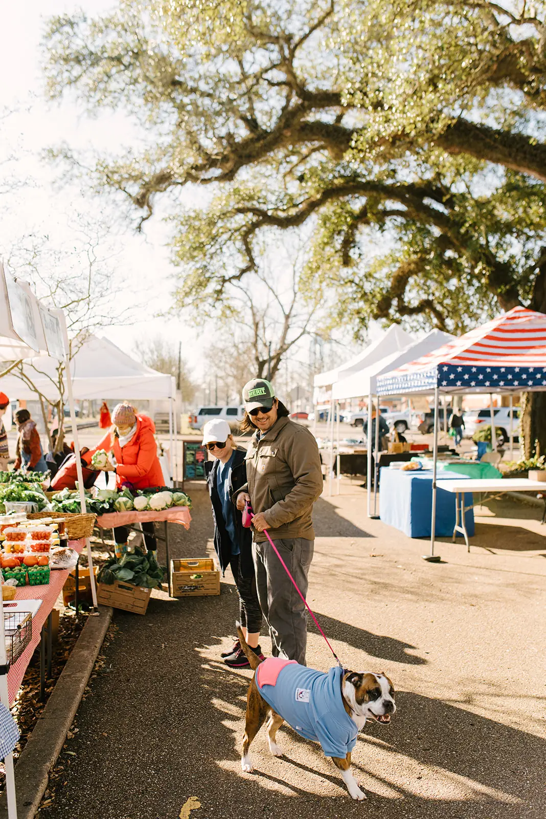 Man and woman at the Hammond Farmers & Artisans Market with a dog wearing a blue and pink jacket on a leash.