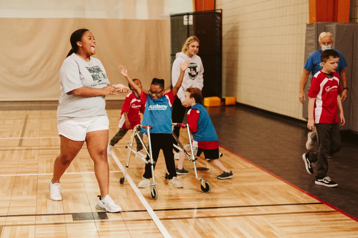 Group of children in the TOPs program and adult supervisors playing and celebrating in an indoor gym, with one child using a walker and raising arms joyfully.