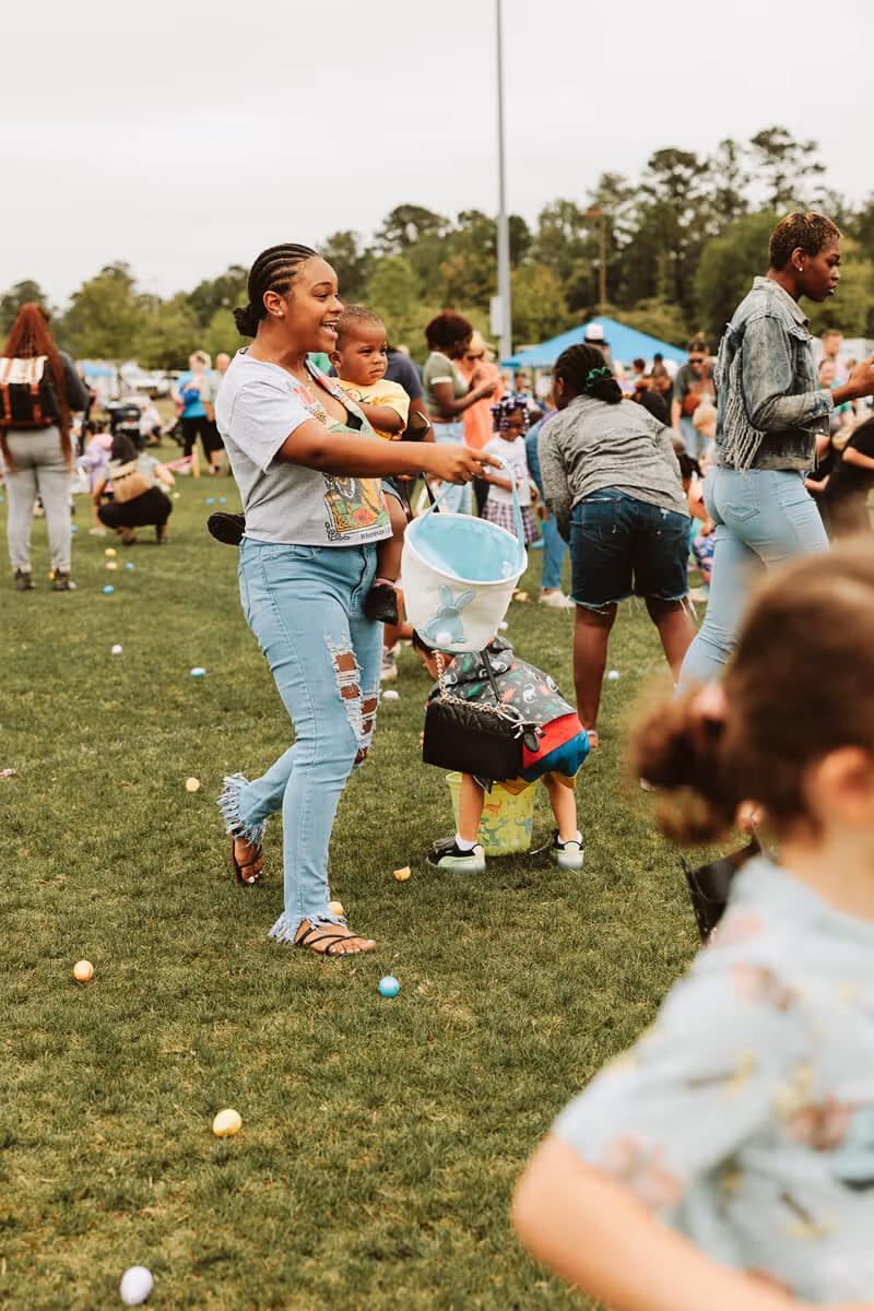 Woman carrying a child and holding a blue bucket during Easter Eggstravaganza egg hunt with scattered colorful eggs on grass.