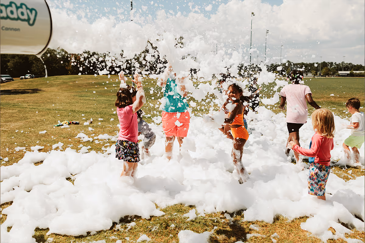 Children playing and laughing in foam bubbles on a sunny grassy field during Back to School Bash.