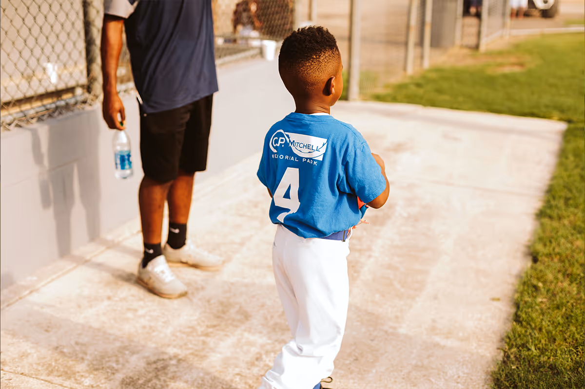 Young baseball player in a blue jersey with C.P. Mitchell branding stands on a pathway holding a ball, with an adult nearby holding a water bottle.
