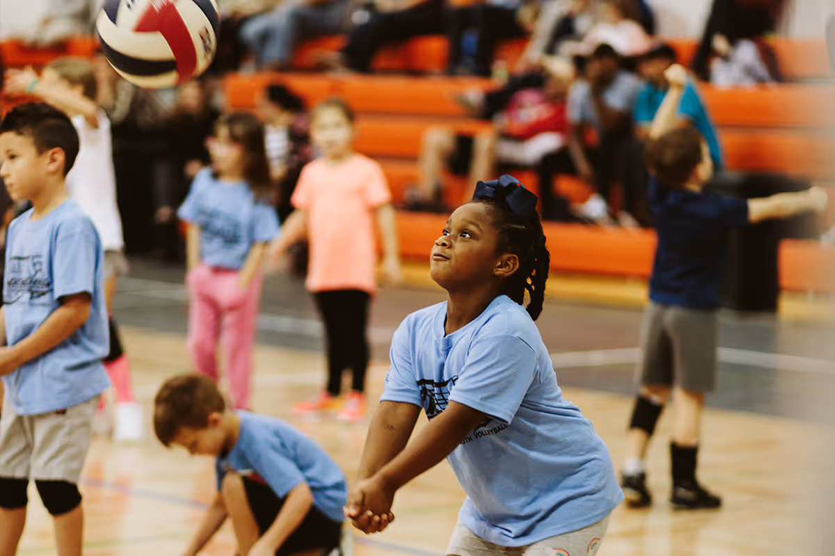 Children playing volleyball indoors, with a girl in a blue shirt preparing to hit the ball.