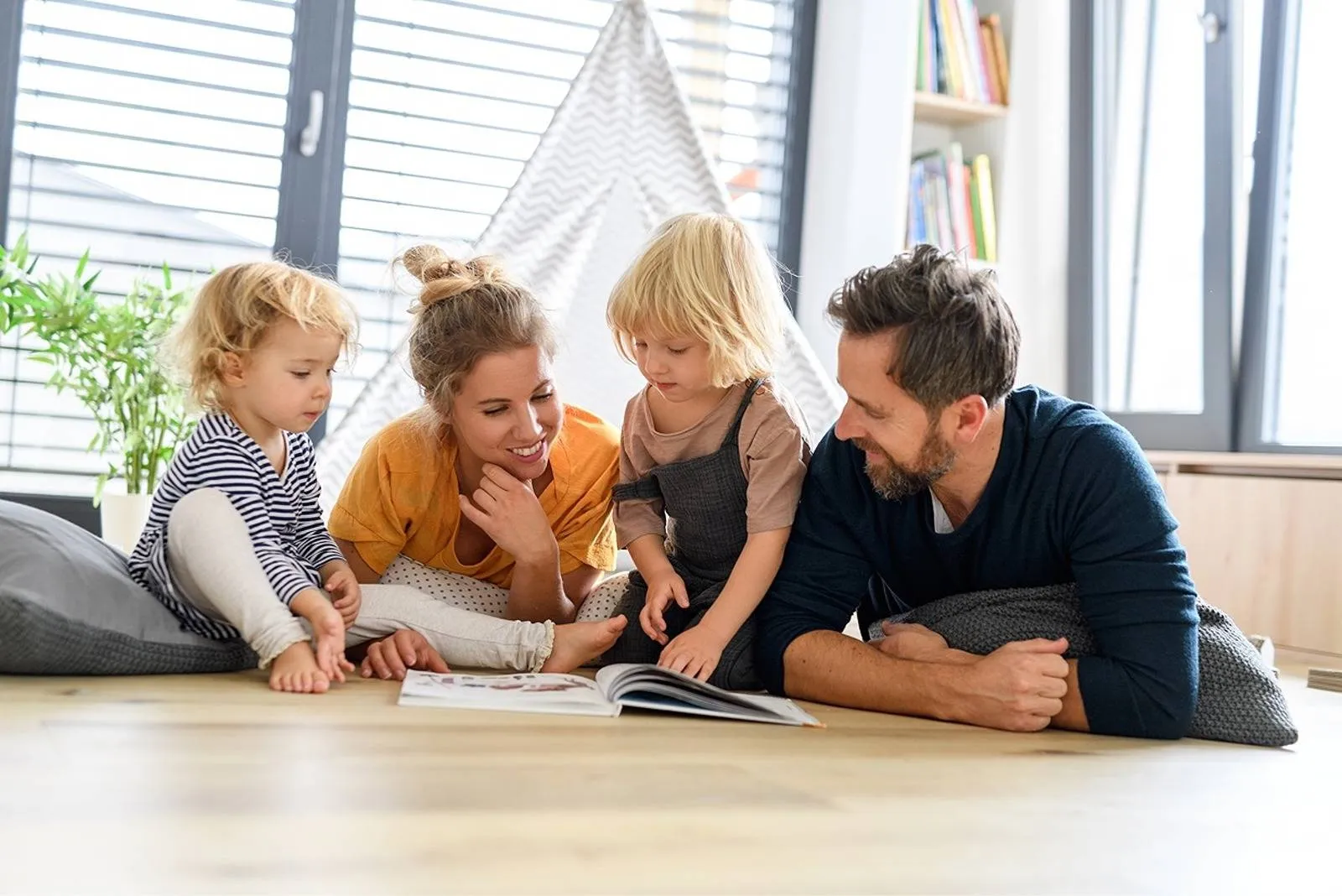 Smiling parents and two young children lying on the floor at home, reading a picture book together near a window with natural light.