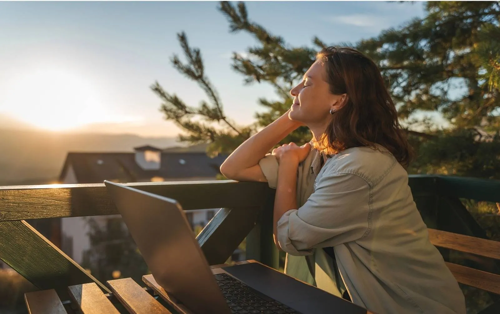 Woman sitting on a balcony with a laptop, smiling peacefully while facing the sunset over rooftops and mountains.