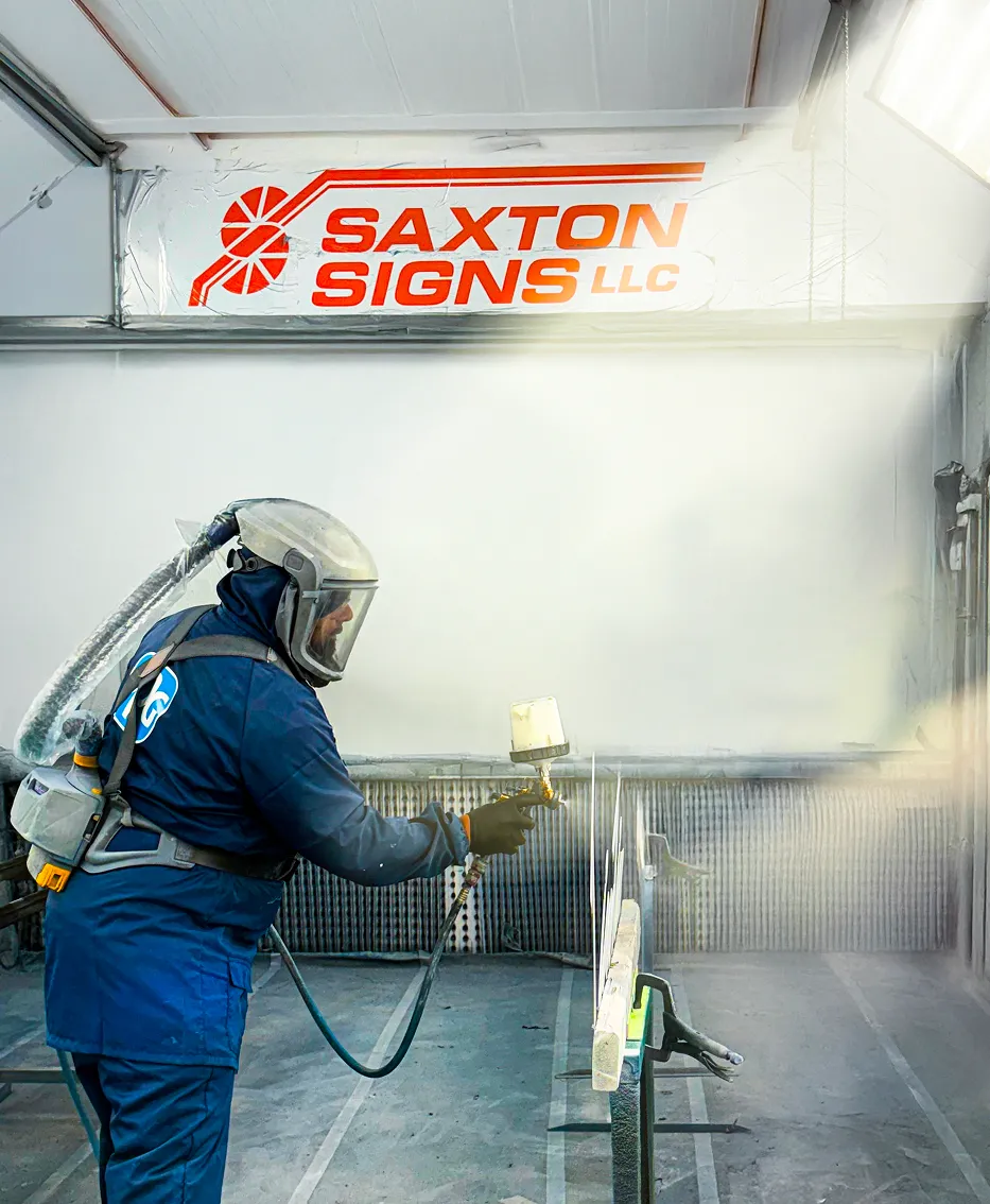 Worker wearing protective gear spray-painting a sign panel inside a booth at Saxton Signs LLC.