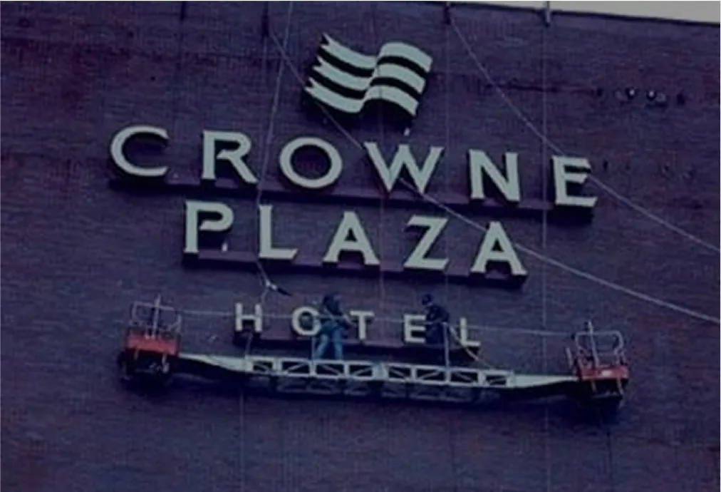 Two workers on suspended platforms installing large signage for Crowne Plaza Hotel on a brick wall.