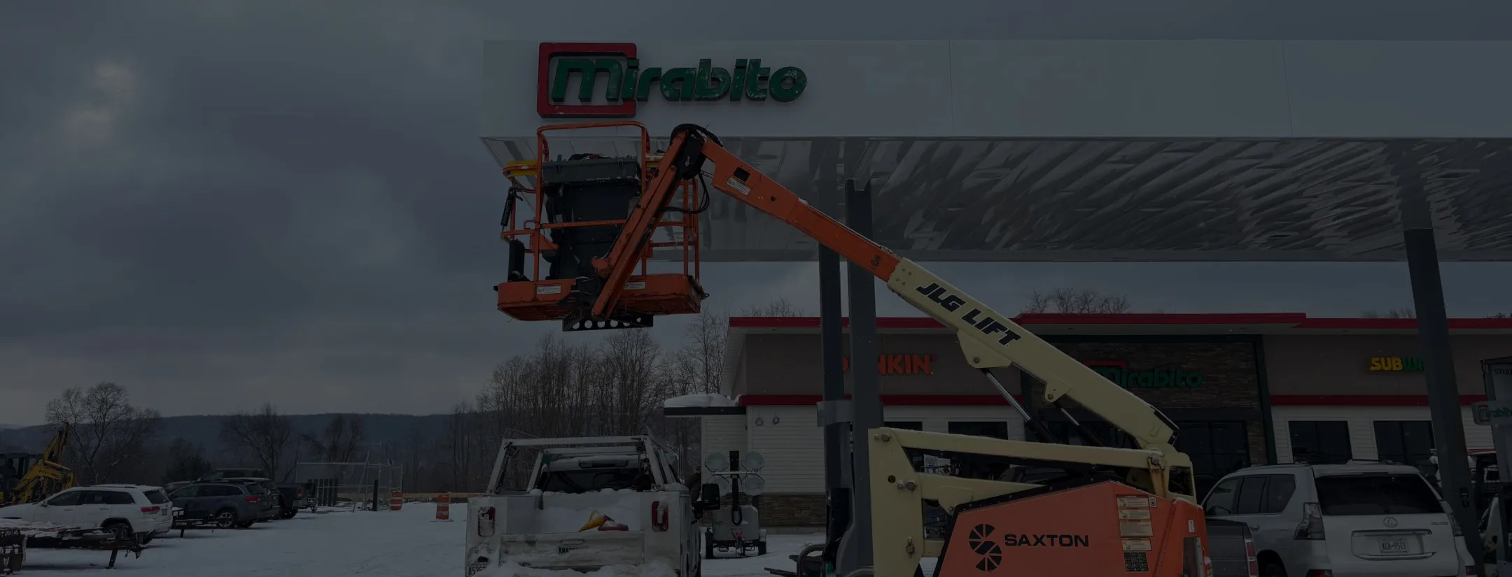 Installation of Mirabito gas station canopy signage using JLG lift, with snowy ground and parked vehicles