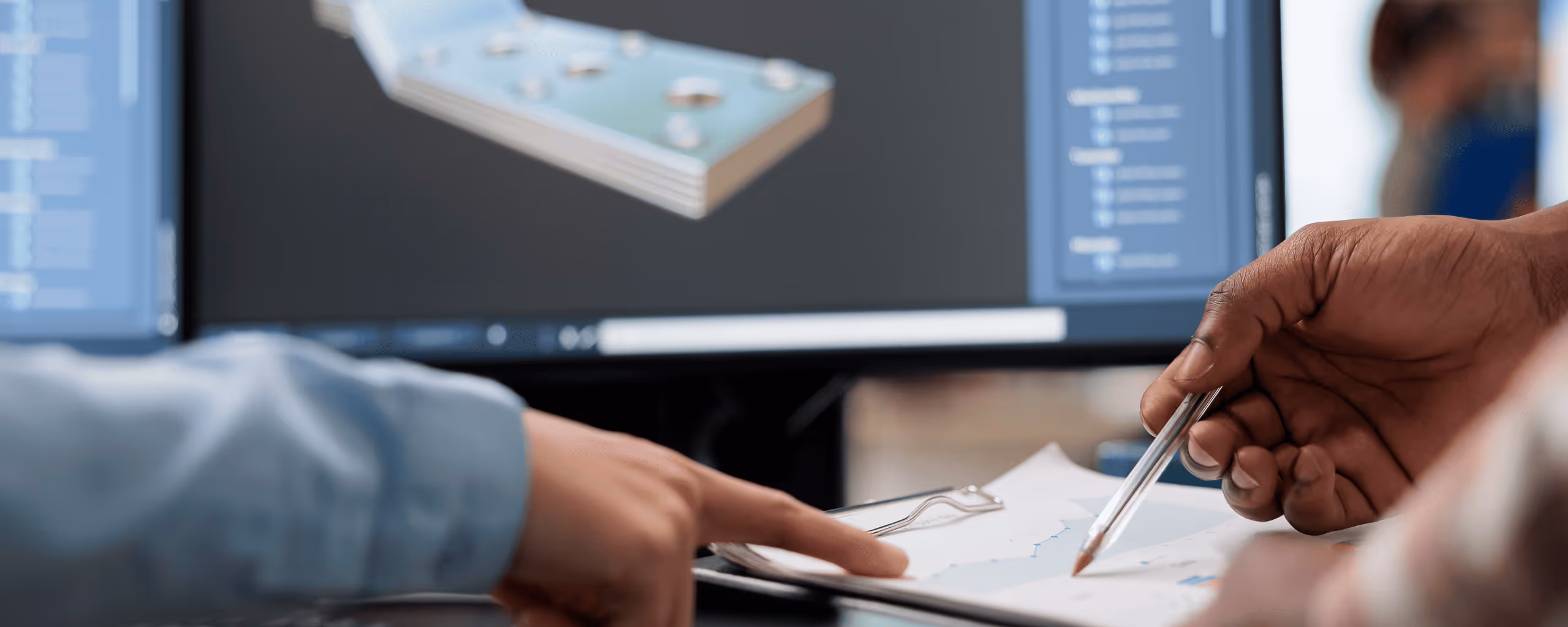 Two people reviewing a technical drawing with a 3D CAD model displayed on a computer screen.