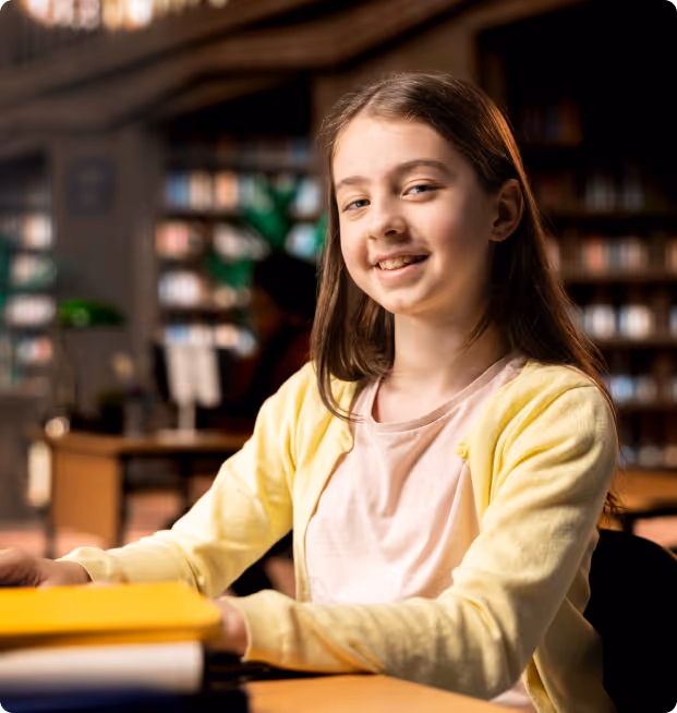 Student in library study area with books stacked on desk, wearing yellow cardigan.