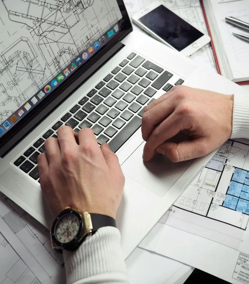 Hands typing on a laptop beside construction drawings