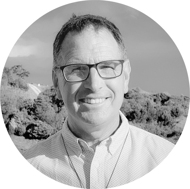 Smiling man with short hair and glasses wearing a buttoned shirt outdoors with trees in the background.