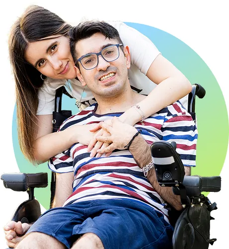 Man in a wheelchair wearing glasses and a striped shirt, embraced and smiling with a woman leaning over him