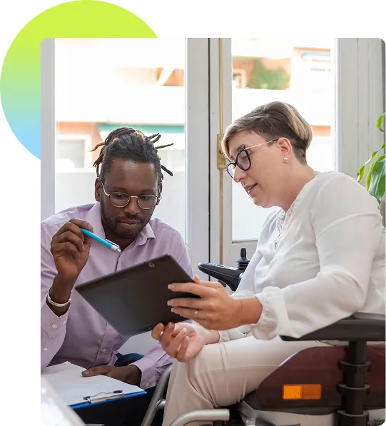 A person in a assistive mobility device talking with someone about something on the tablet screen they're holding