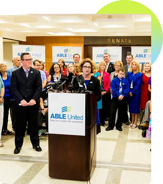 Jenn Sikora at a podium in front of a Senate sign and a crowd of people, speaking on behalf of ABLE United's launch