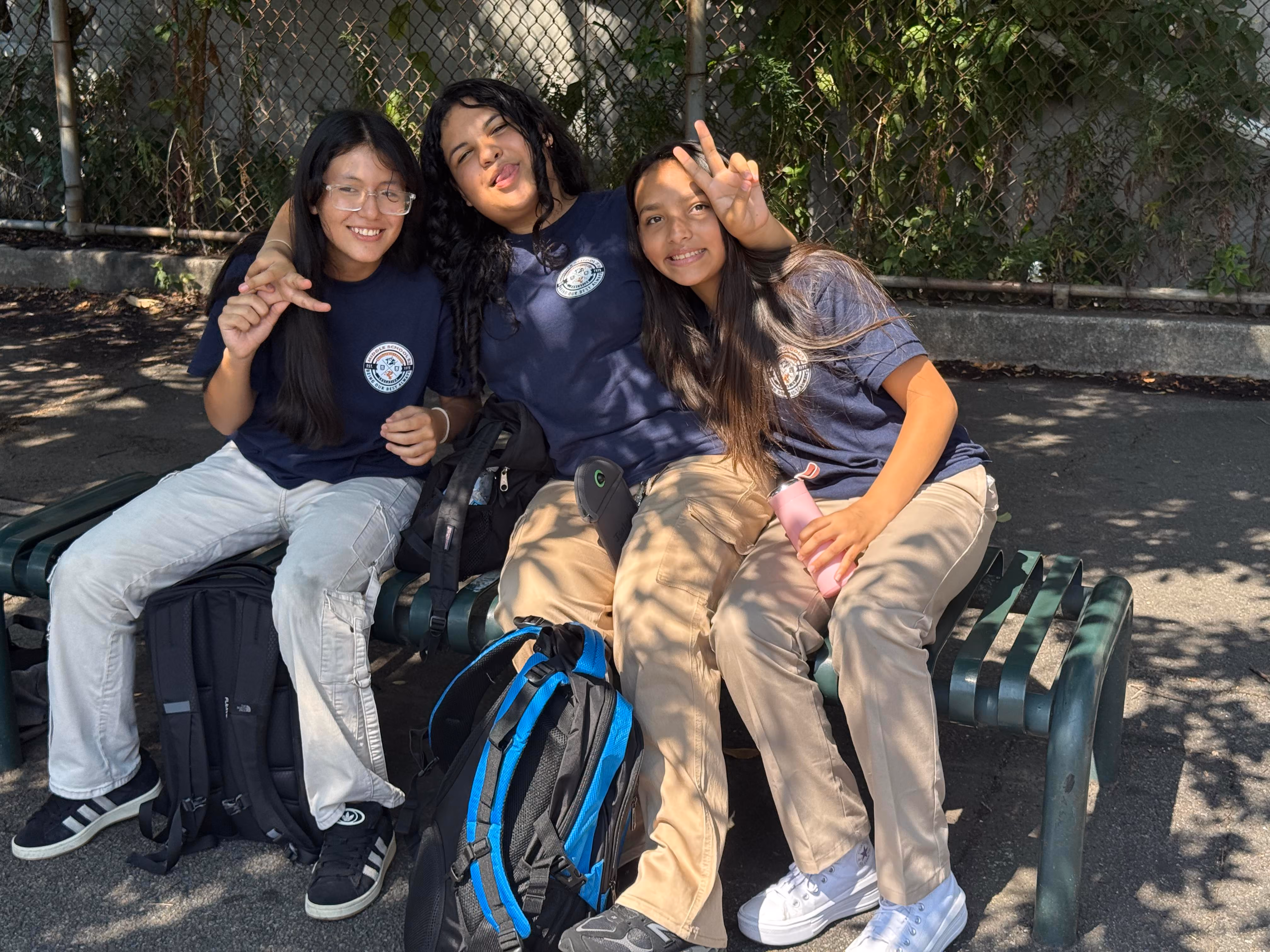 Three students in school uniforms sitting on outdoor bench smiling