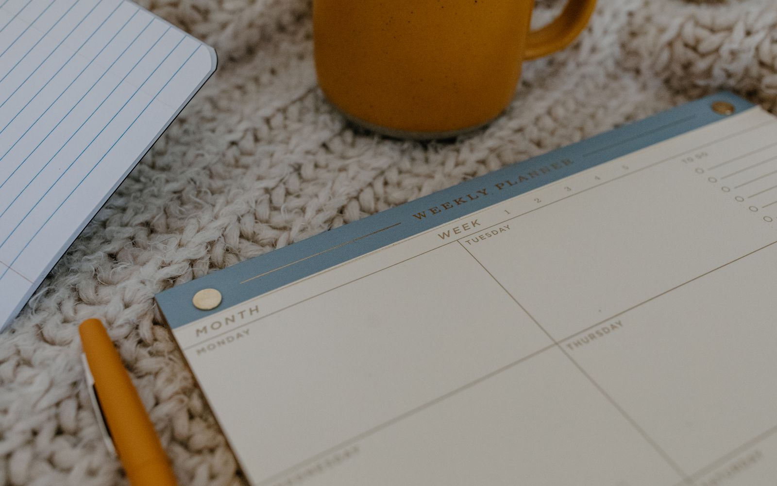 An undated weekly planner, pen, notebook and yellow mug on knitted fabric.