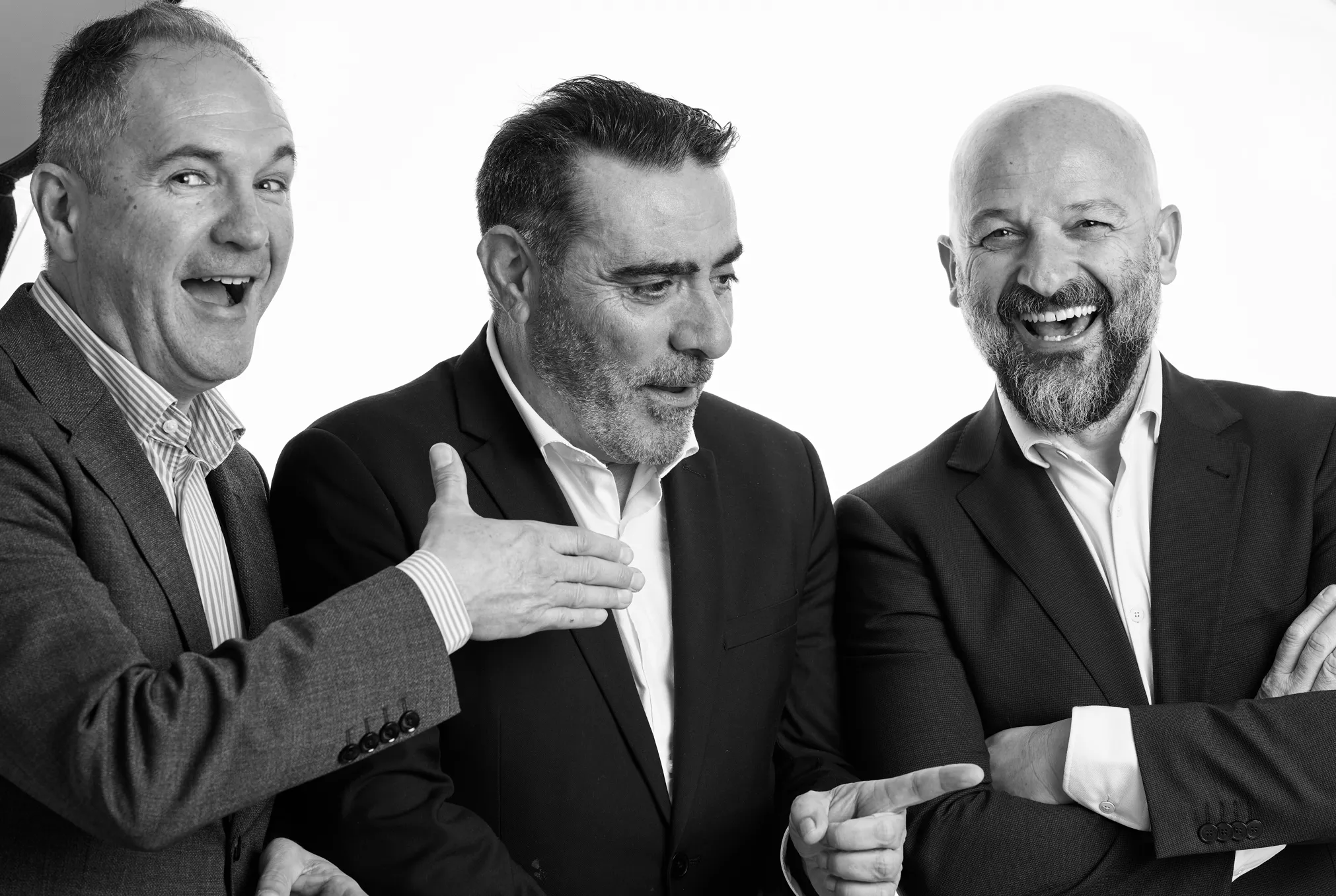 Tres hombres de negocios sonrientes y vestidos formalmente en blanco y negro, dos señalando al hombre de la derecha que ríe con los brazos cruzados.