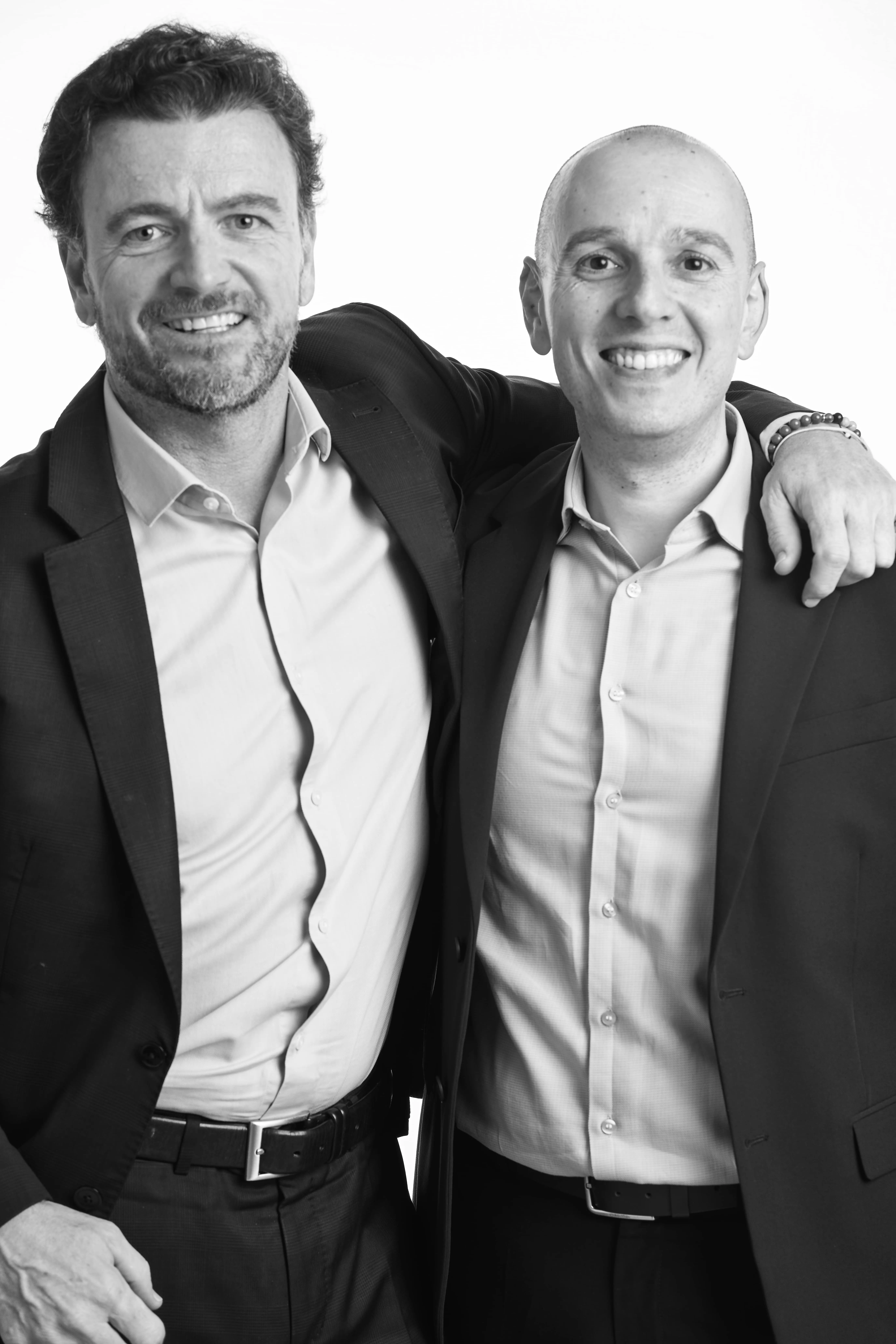 Dos hombres sonrientes con trajes oscuros y camisas claras, uno con el brazo sobre el hombro del otro.