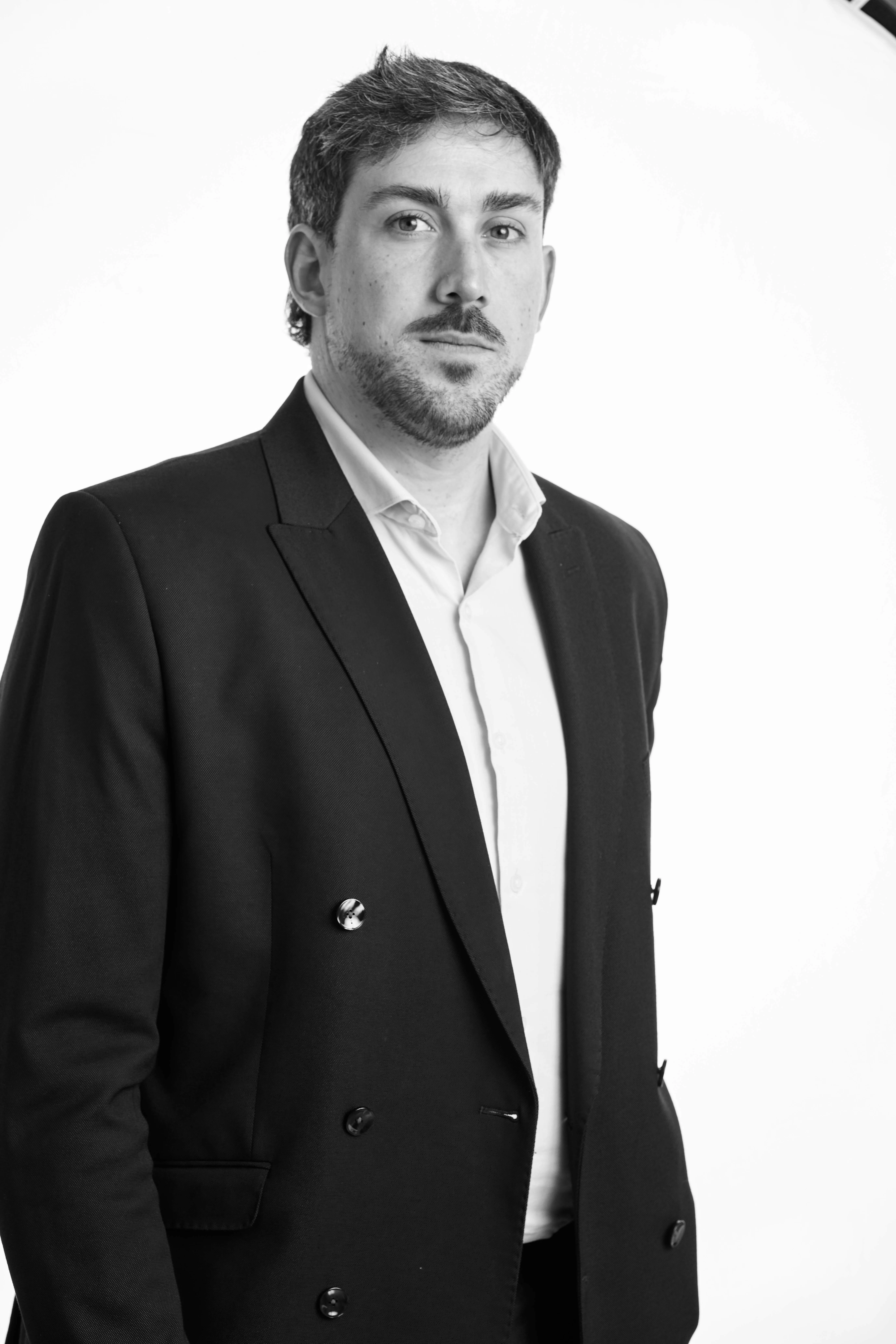 Hombre con barba y bigote vestido con blazer oscuro y camisa blanca sobre fondo blanco.