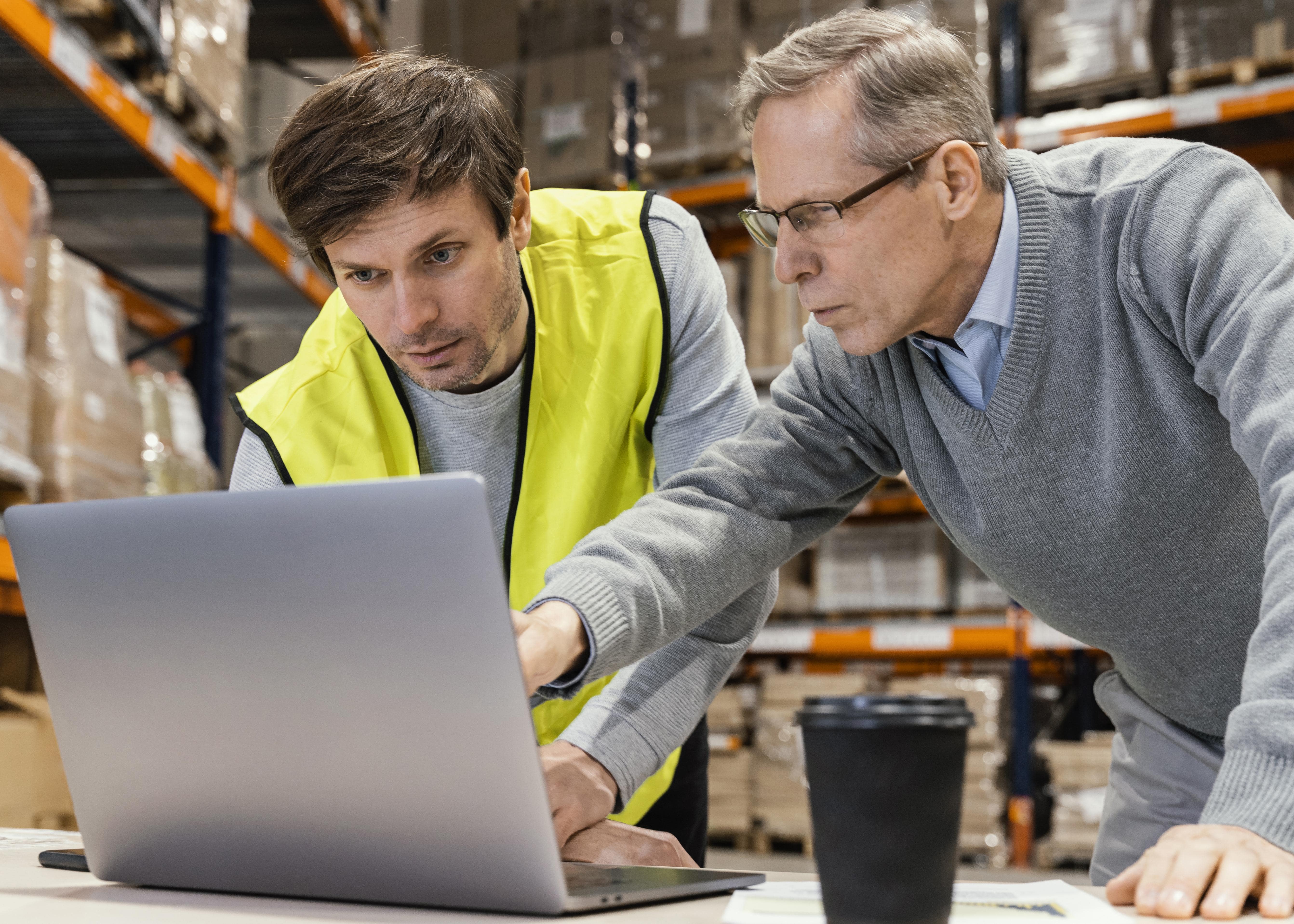 Dos hombres en un almacén revisan la información en una computadora portátil sobre una mesa con un vaso de café.