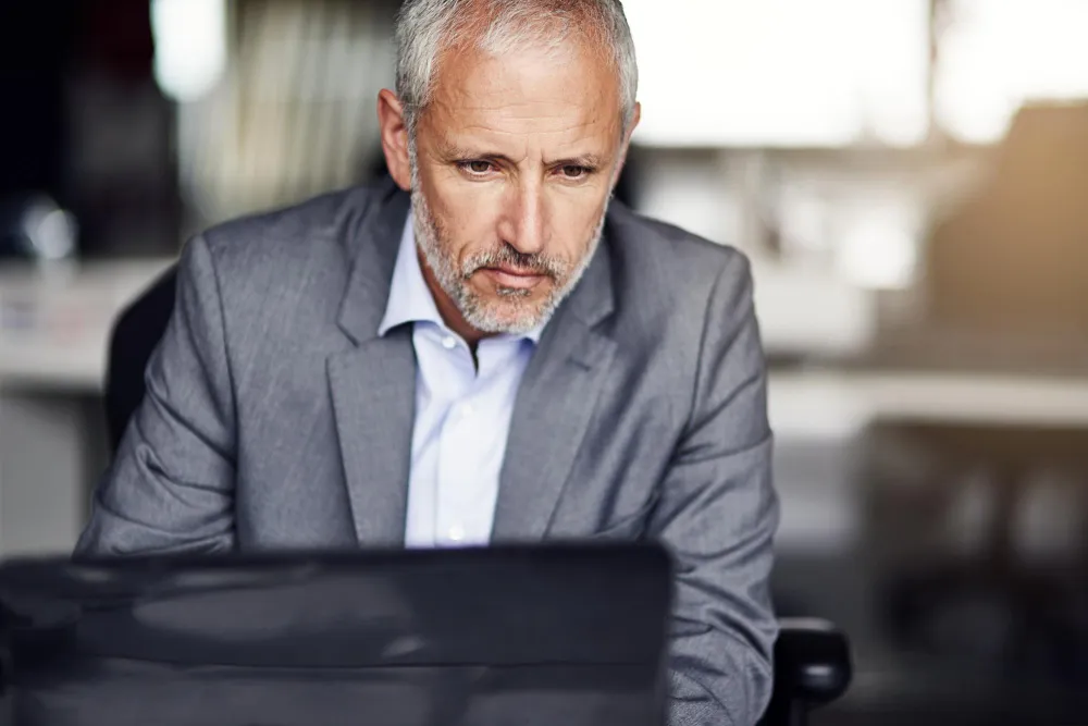 Hombre de mediana edad con chaqueta gris y camisa blanca mirando seriamente una computadora portátil en una oficina.