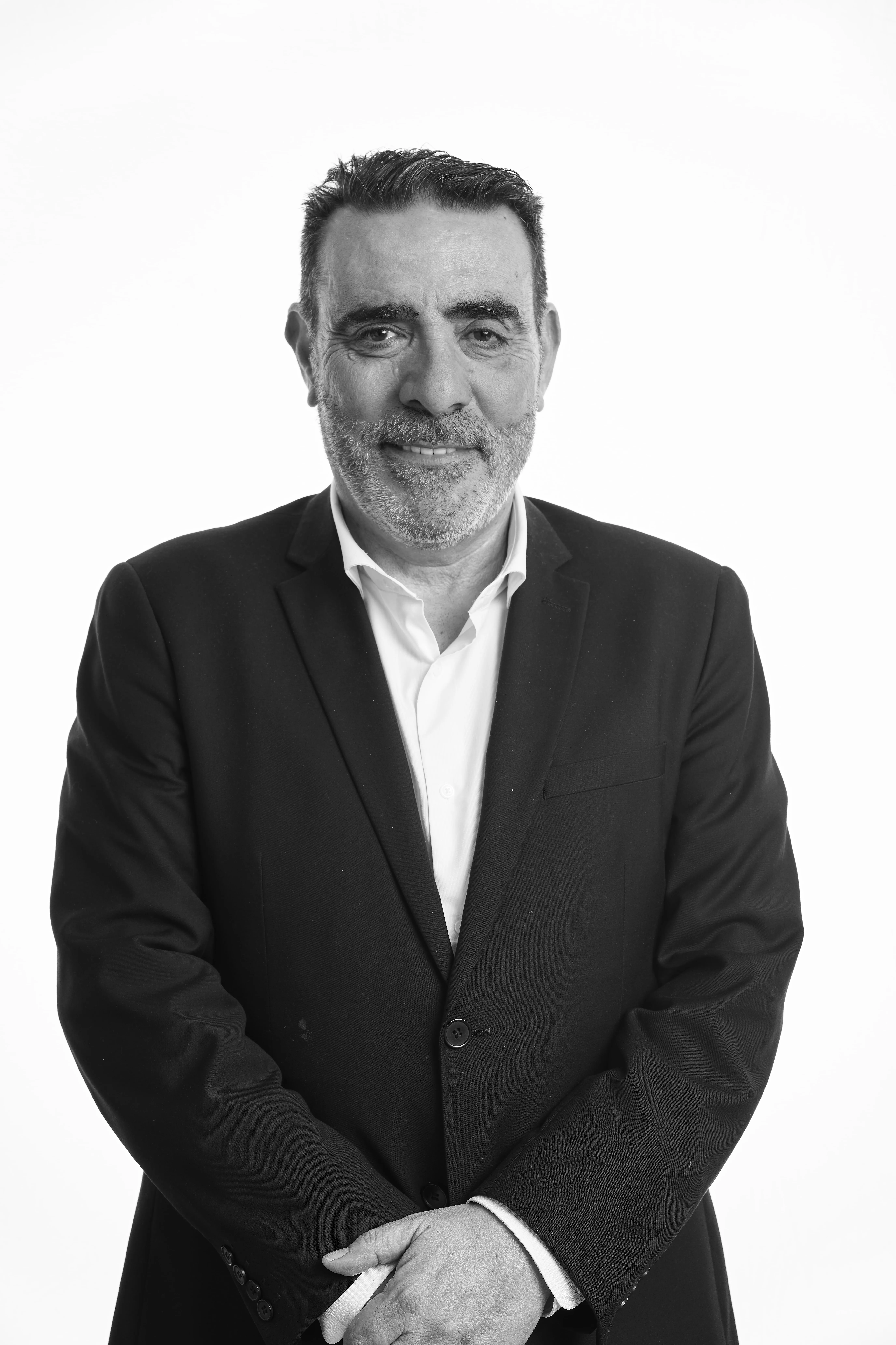 Hombre de mediana edad con barba y cabello corto, vestido con traje oscuro y camisa blanca, posando con las manos cruzadas.