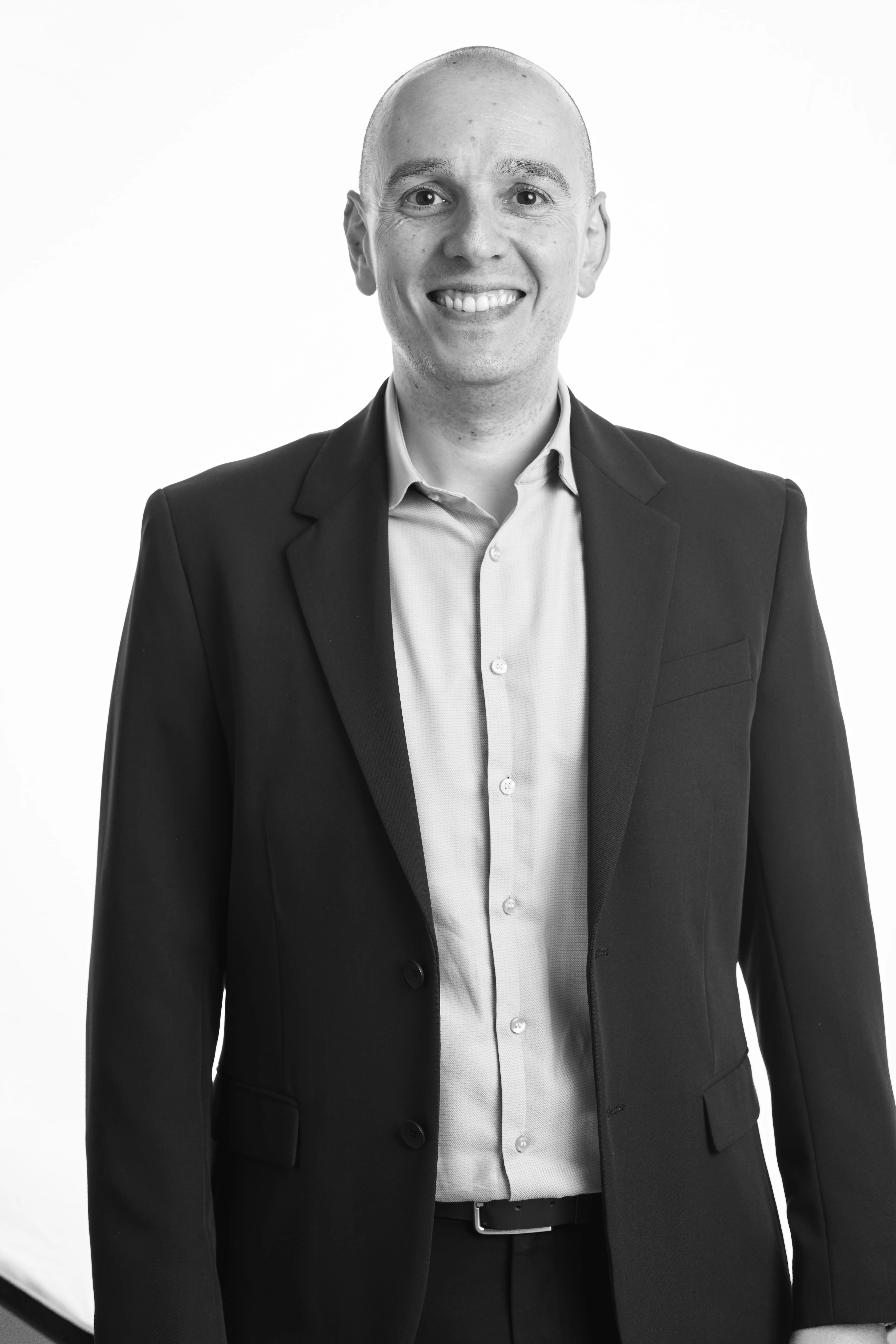 Hombre calvo sonriendo, vistiendo traje oscuro y camisa clara, posando sobre fondo blanco.