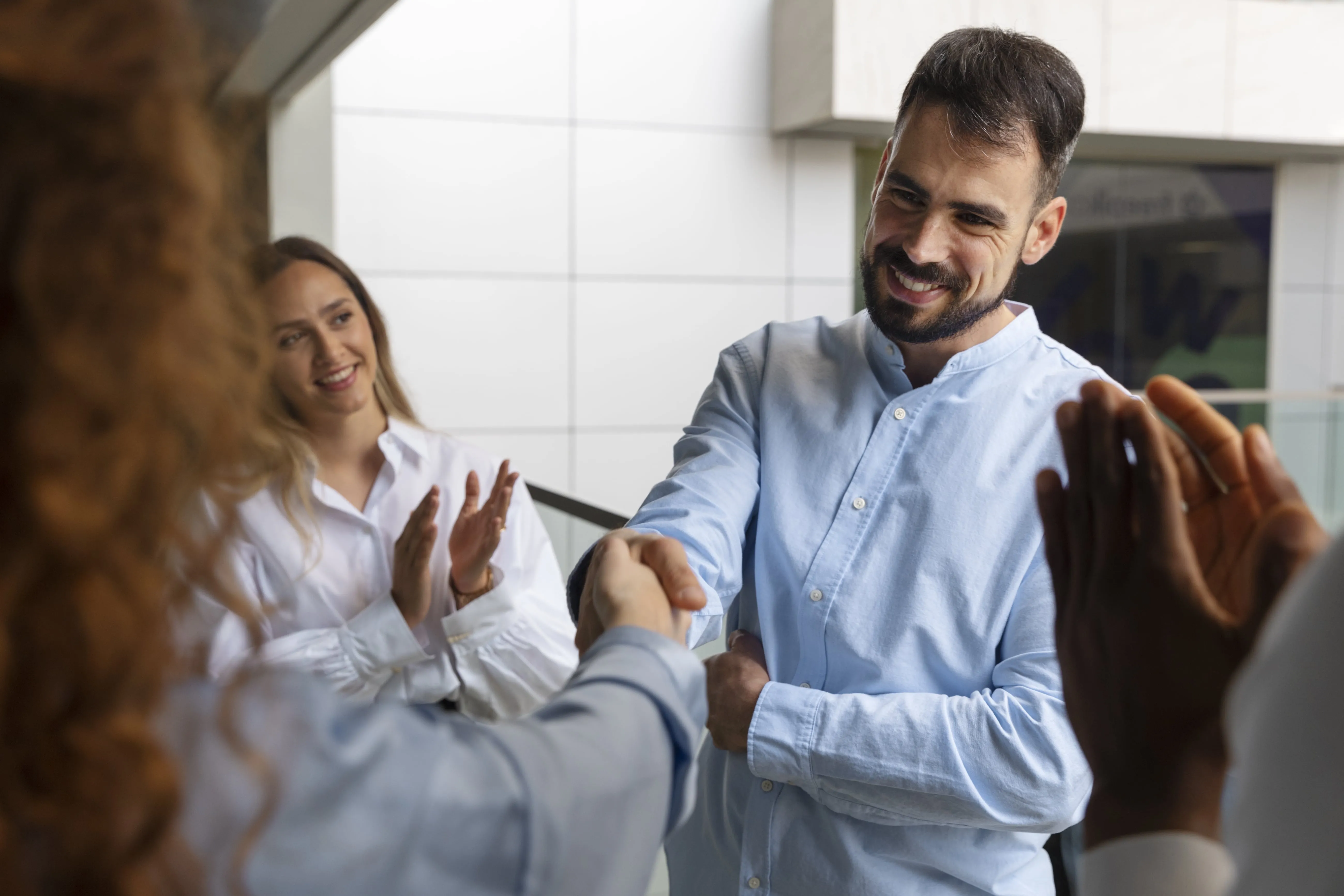 Hombre sonriente dándose la mano con alguien mientras otra persona aplaude en un entorno de oficina moderno.