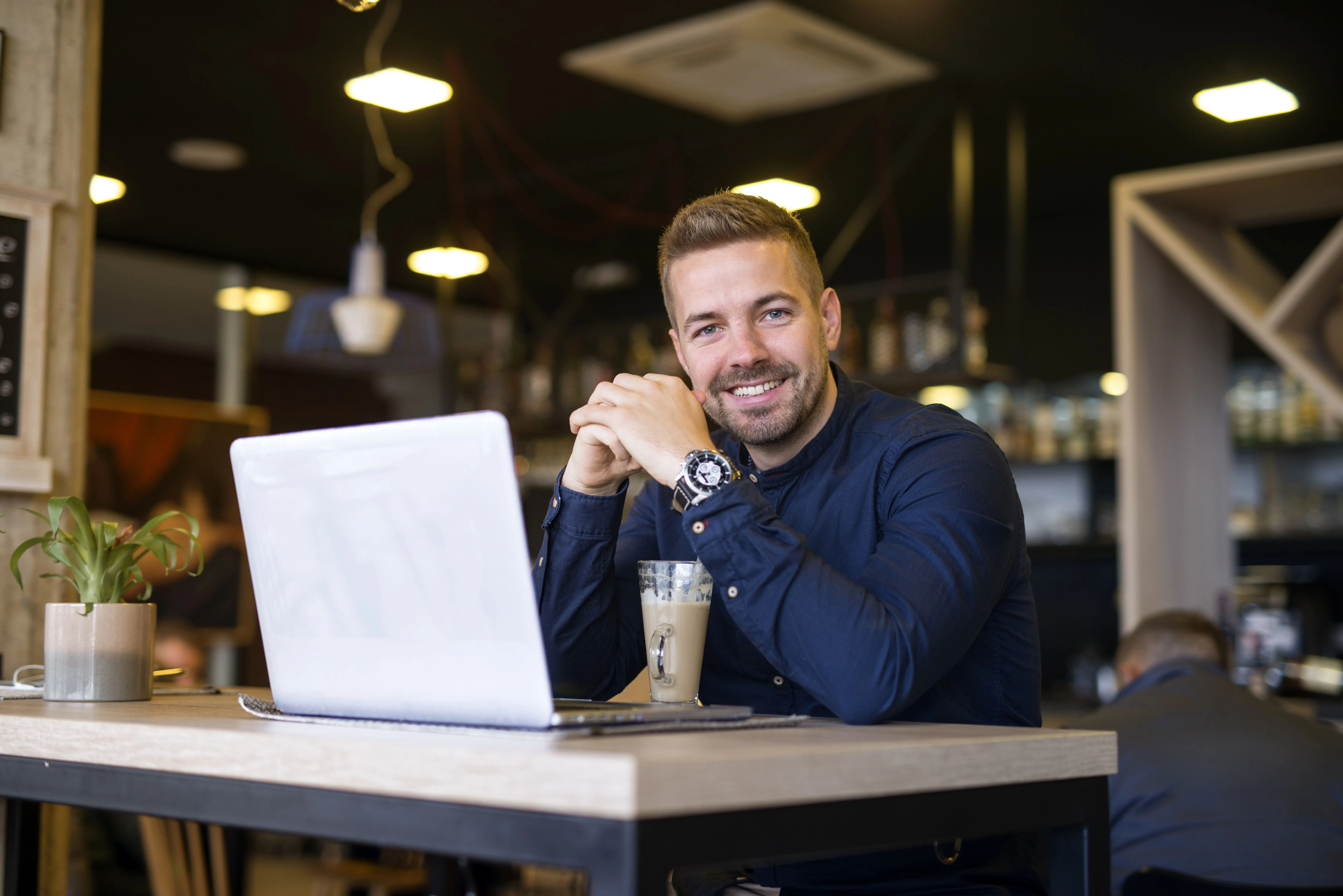 Hombre sonriente sentado en una mesa con una laptop y un café en una cafetería moderna.