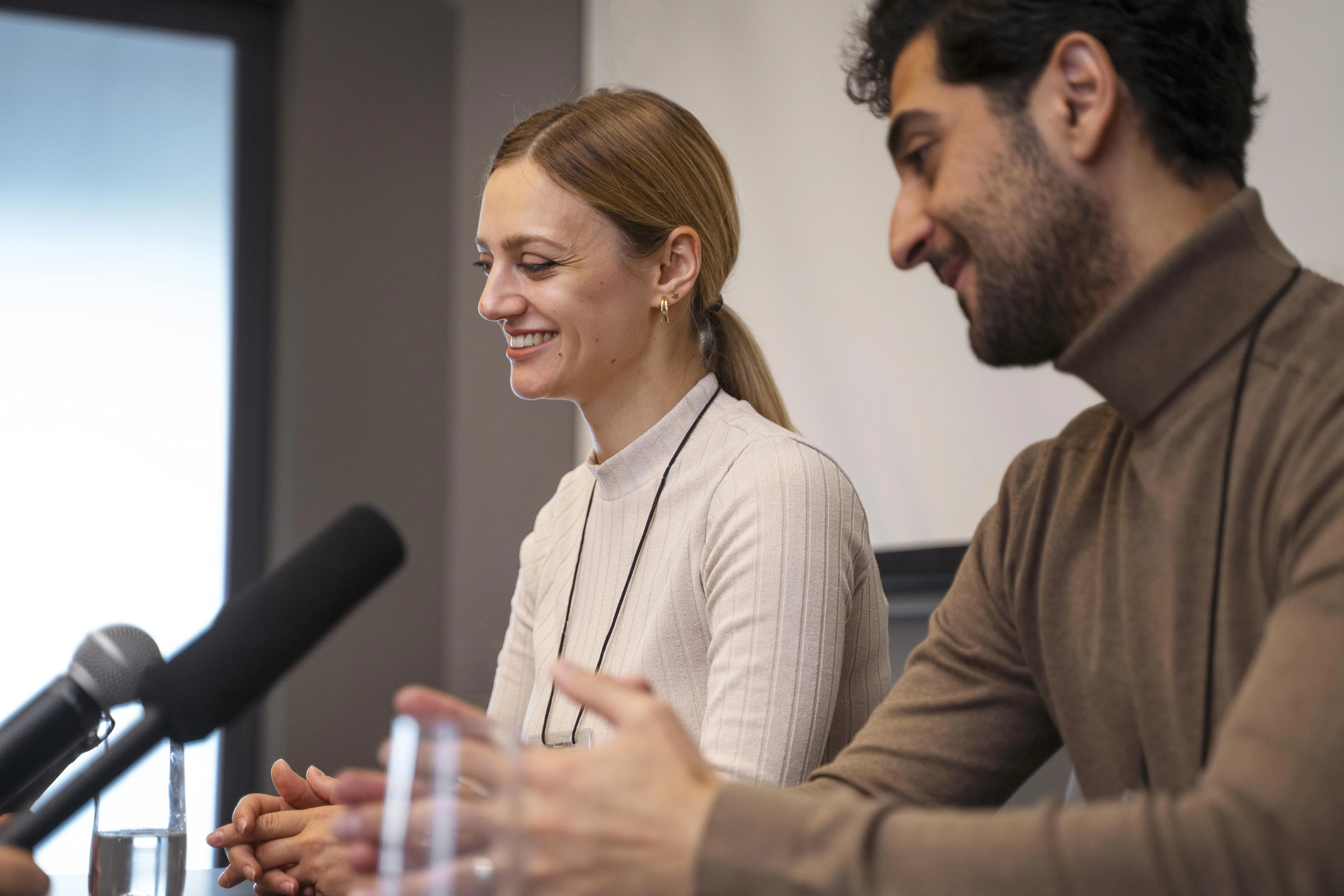 Dos personas sonrientes sentadas frente a micrófonos en una conferencia o entrevista.