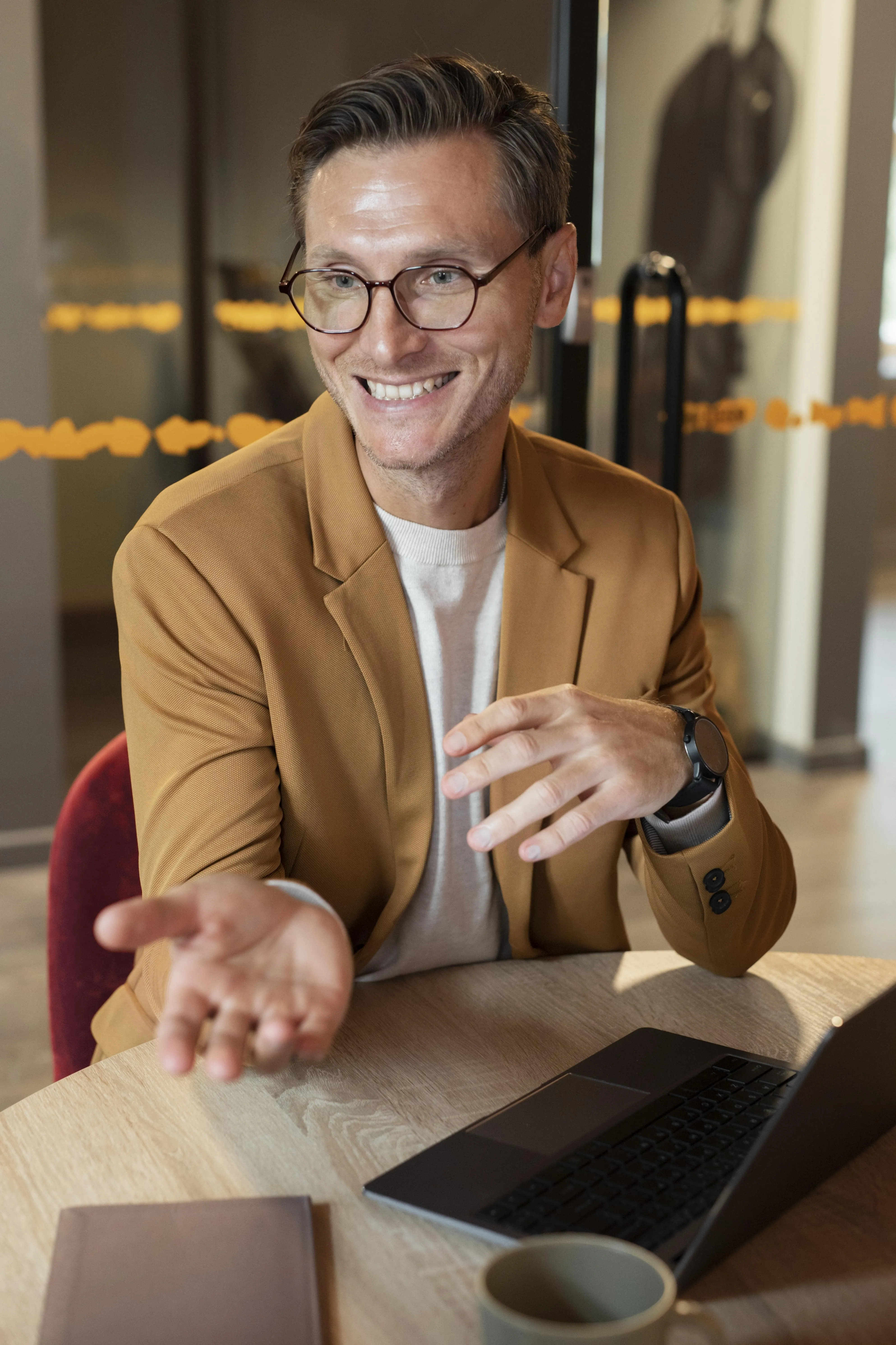 Hombre sonriente con gafas y chaqueta marrón hablando y gesticulando frente a una laptop en una mesa.
