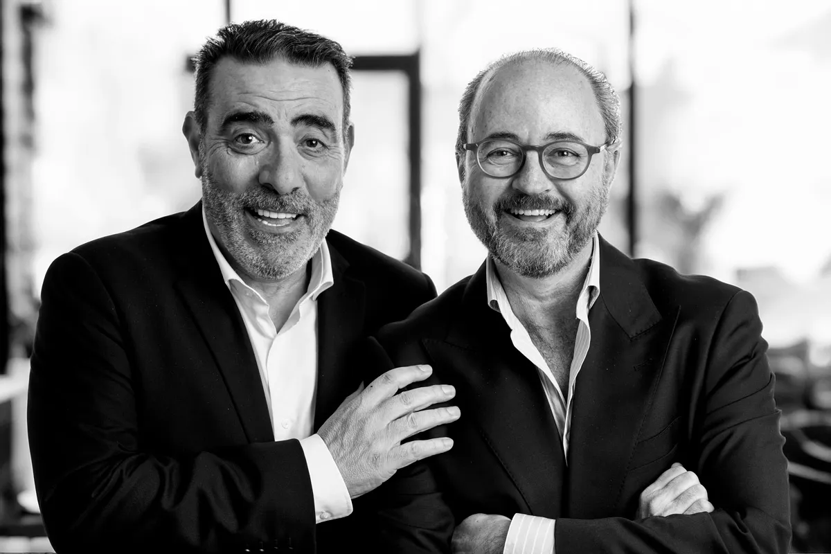 Dos hombres sonrientes con trajes oscuros, uno con gafas, posan juntos en un ambiente interior.