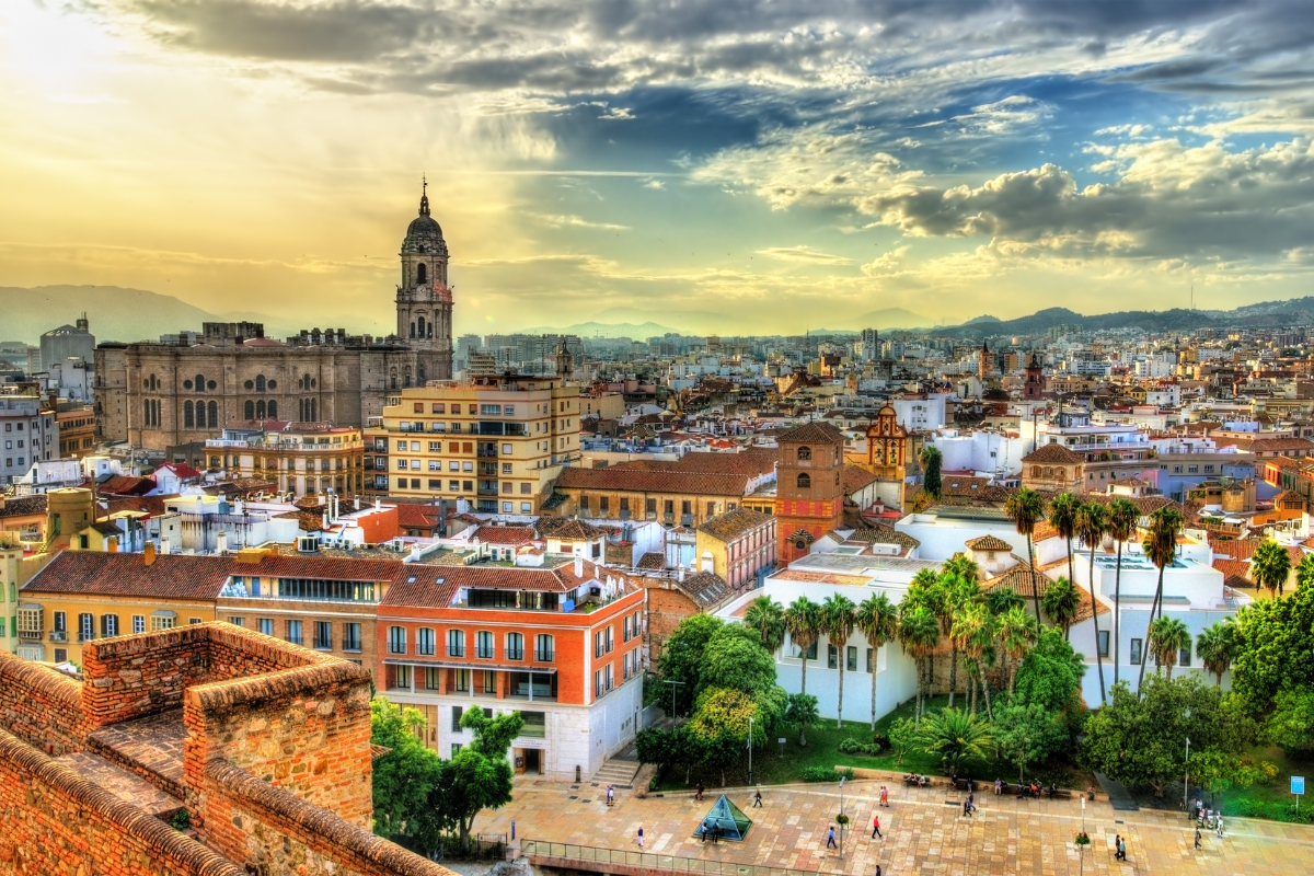 Vista panorámica de Málaga con la catedral al atardecer, edificios variados y árboles en primer plano.
