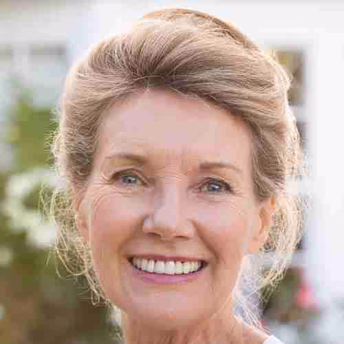 Retrato de una mujer mayor sonriendo con cabello rubio recogido y fondo desenfocado de jardín.