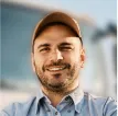 Hombre sonriente con gorra marrón y camisa azul claro de trabajo.