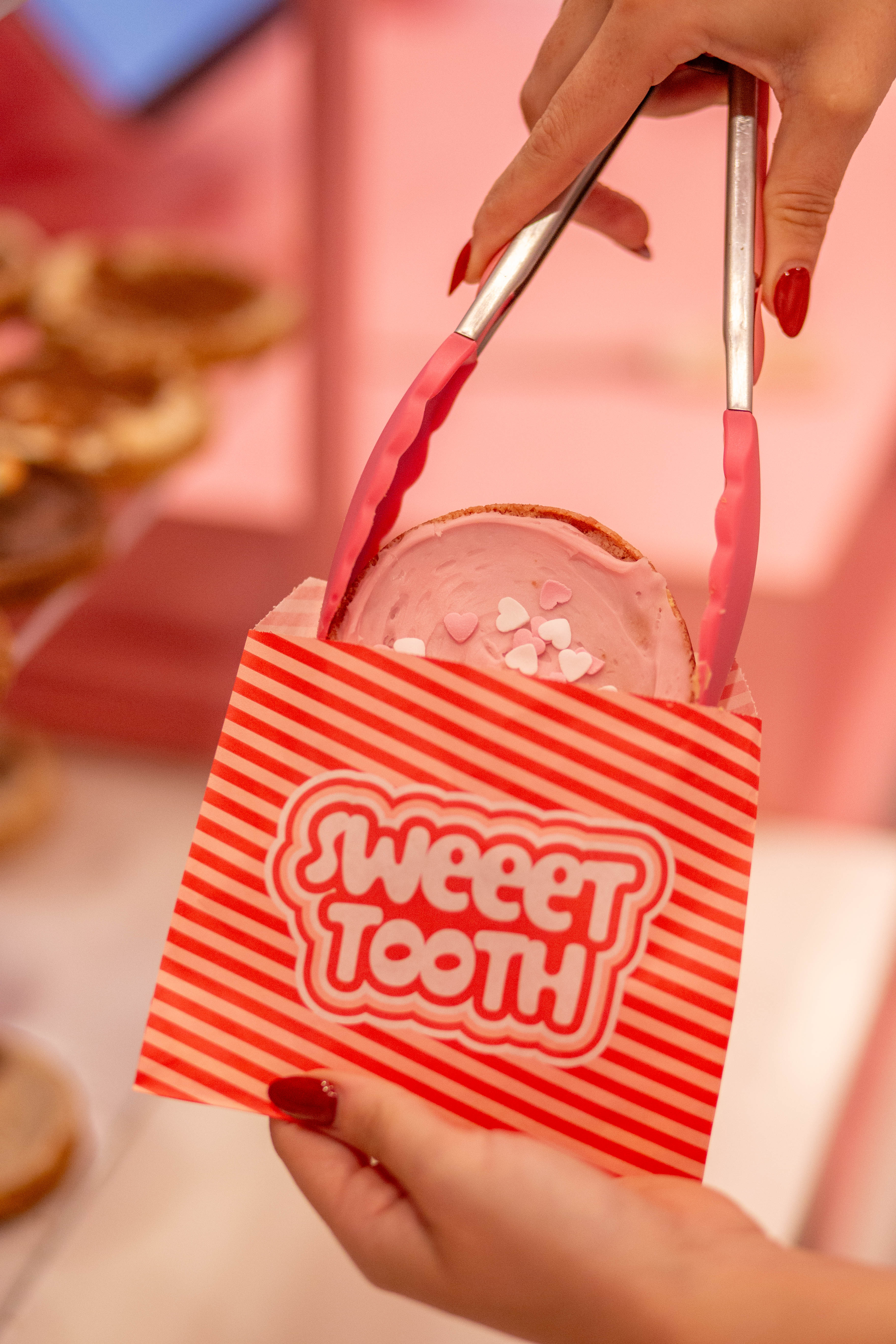 Hands with red nail polish using pink tongs to place a pink frosted cookie with heart sprinkles into a red and white striped 'Sweeet Tooth' bag.