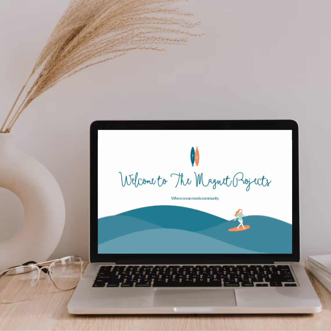 Laptop on wooden desk displaying a website homepage titled The Magnet Projects with an ocean wave graphic and a surfer illustration.
