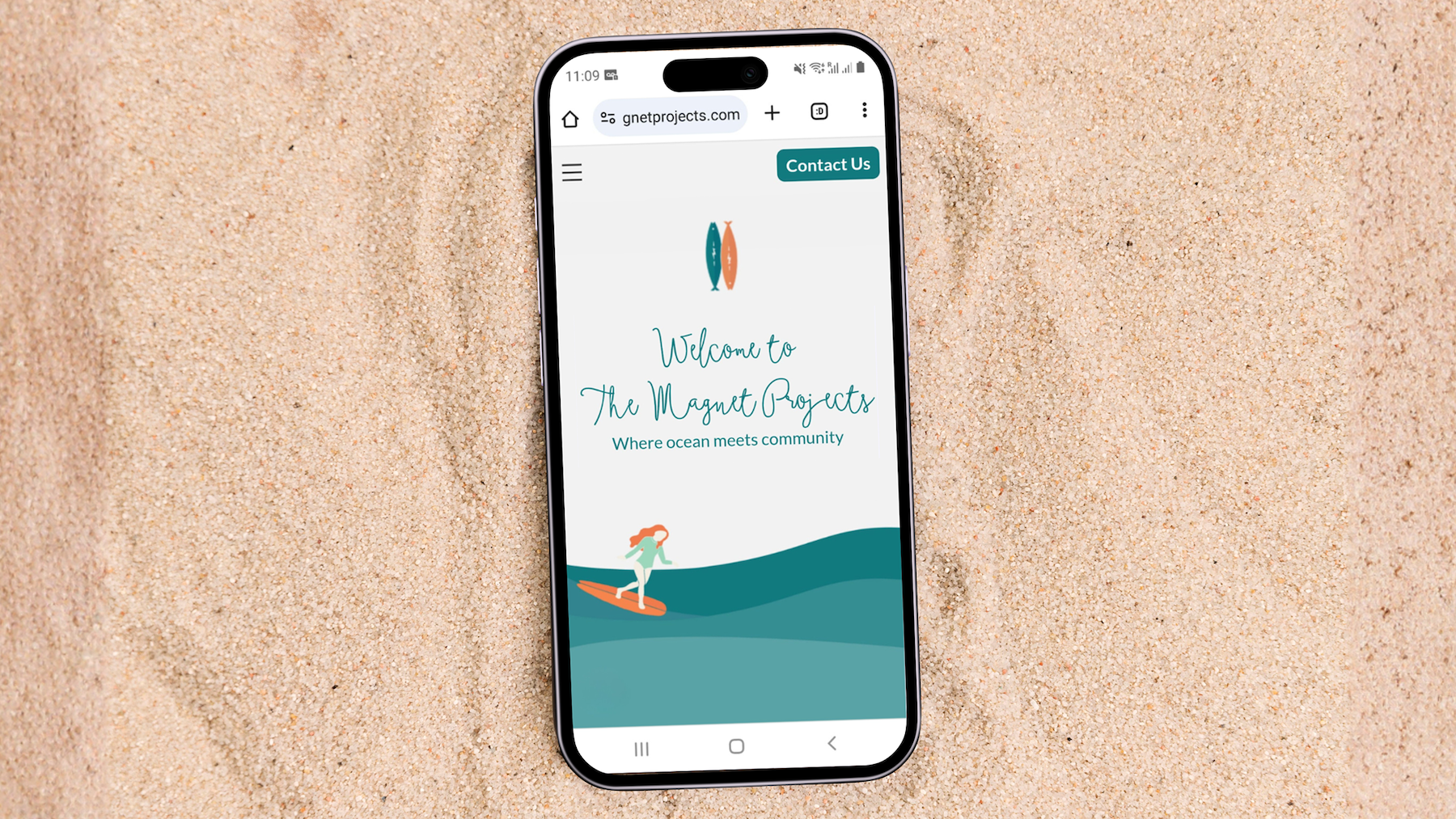 Smartphone on sand showing website with logo of two fish, text 'Welcome to The Magnet Projects' and a surfer illustration on teal waves.