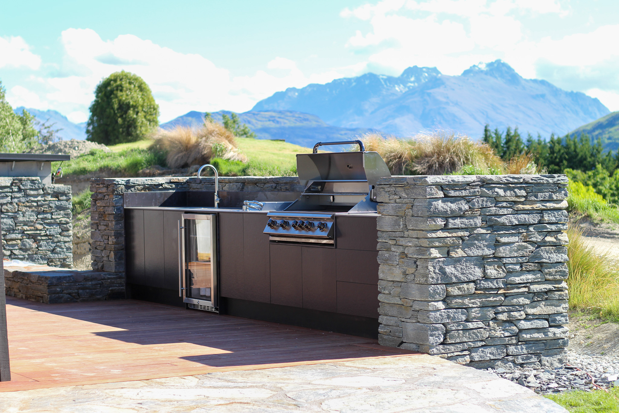 Outdoor kitchen with a stainless steel grill, sink, and mini fridge set in a stone counter with mountains in the background.