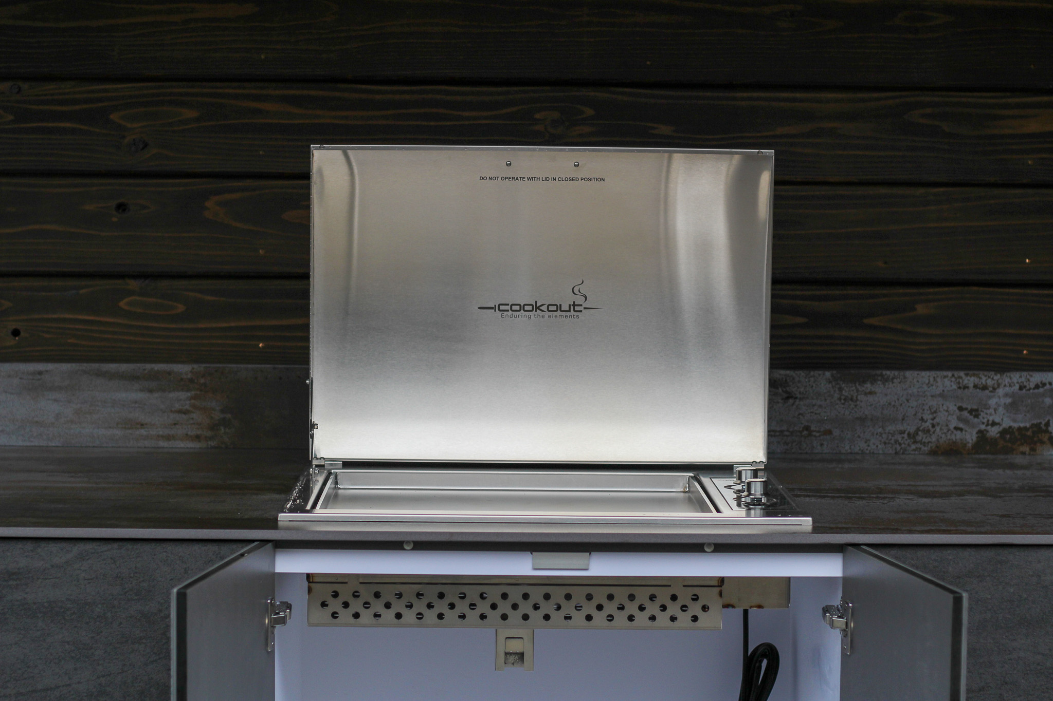 Stainless steel outdoor grill with lid open and two control knobs on the right, set against a dark wooden wall.