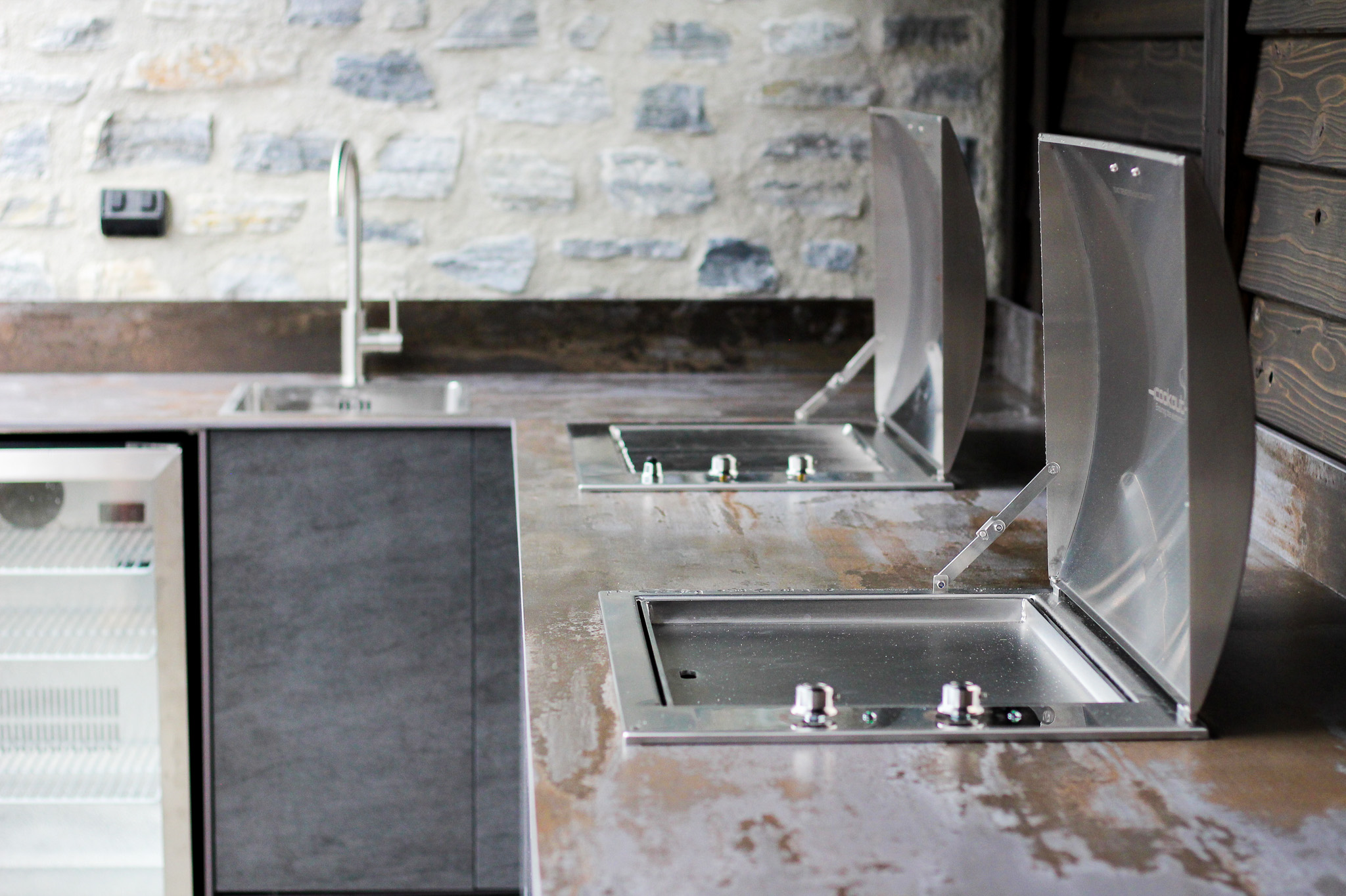 Modern outdoor kitchen countertop with two built-in stainless steel grills and a sink against a stone wall.