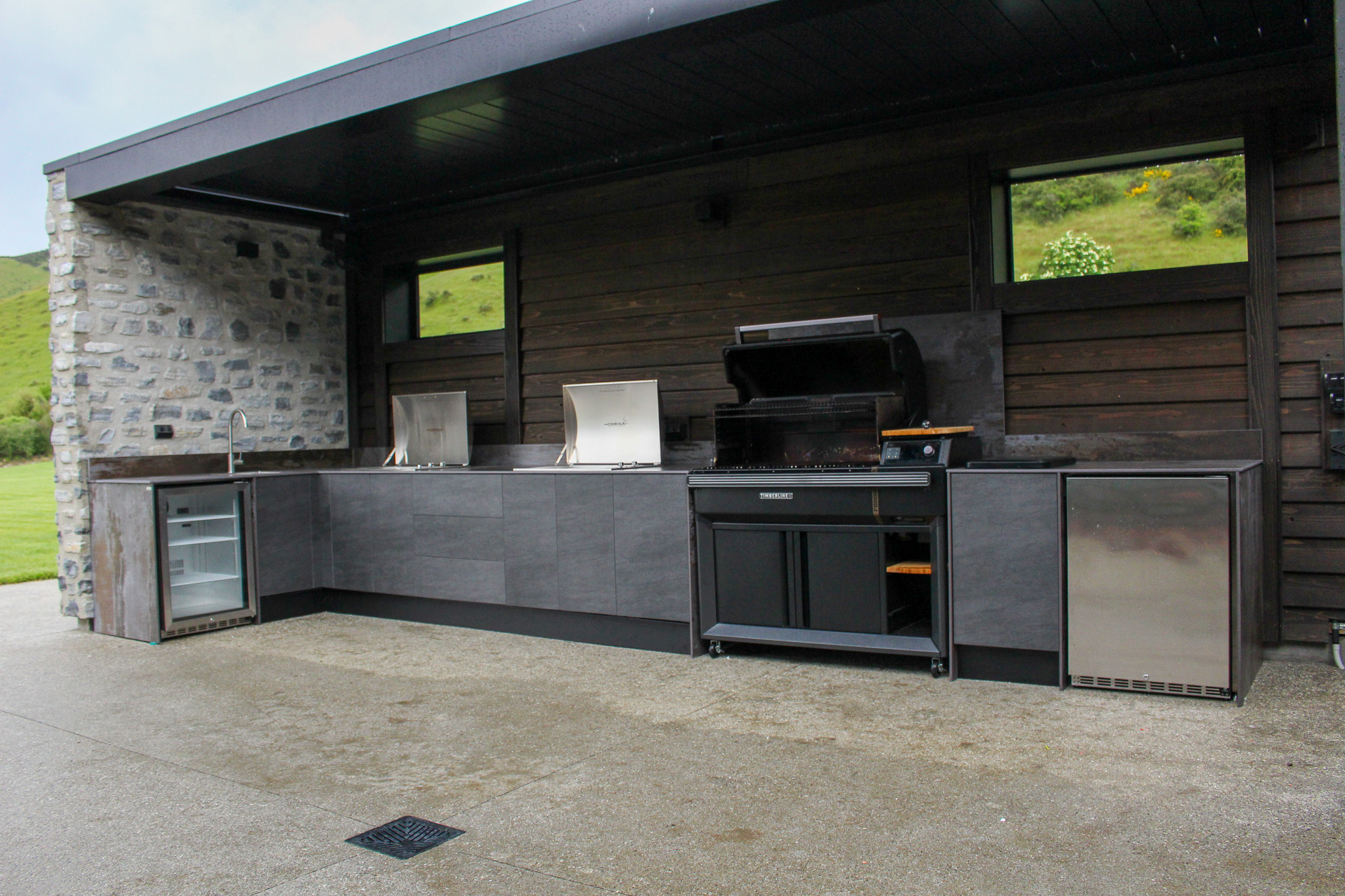 Modern outdoor kitchen with built-in sink, two covered cooktops, a large barbecue grill, and two mini fridges set against a wooden and stone wall.