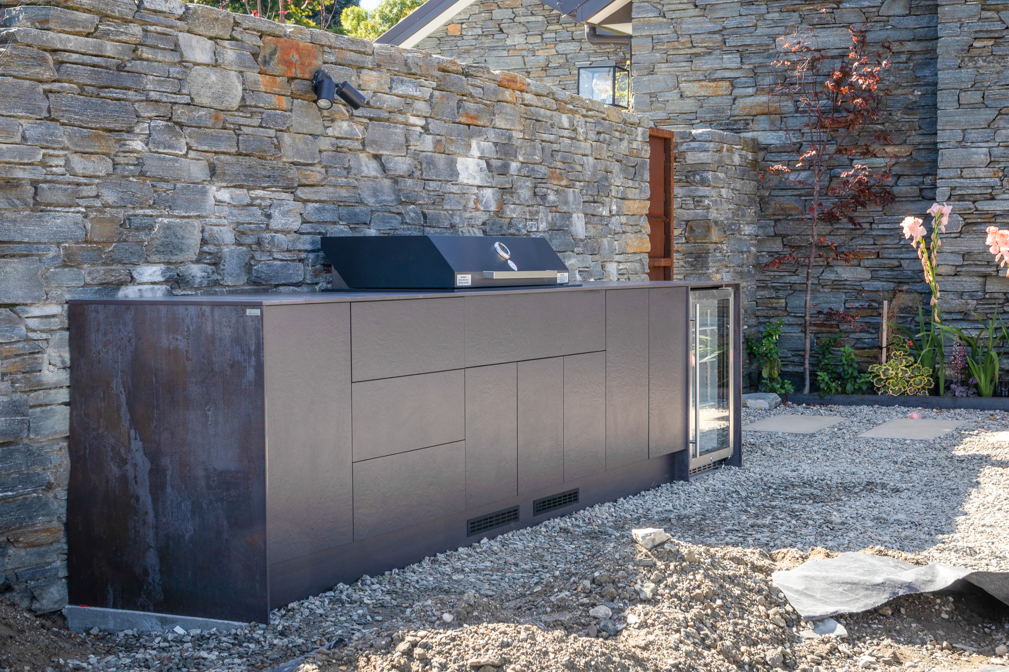 Outdoor kitchen setup with a built-in black grill and glass-door fridge against a stone wall and gravel ground.