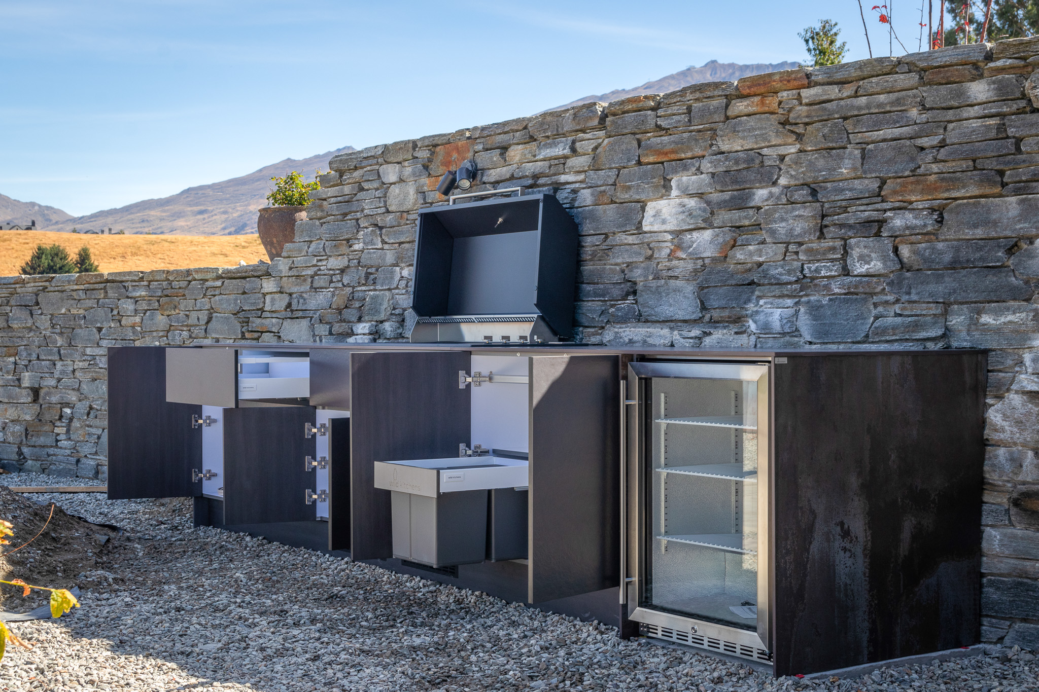 Outdoor kitchen with black cabinets, built-in grill, sink, and glass-door refrigerator against a stone wall.
