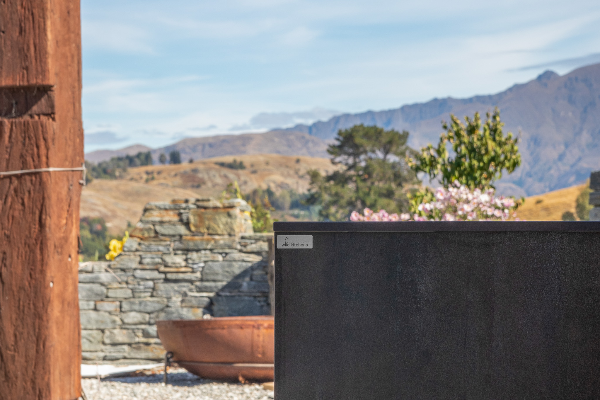 Outdoor kitchen area with a dark metal panel labelled Wild Kitchens, stone wall, fire pit, and mountains in the background under a blue sky.