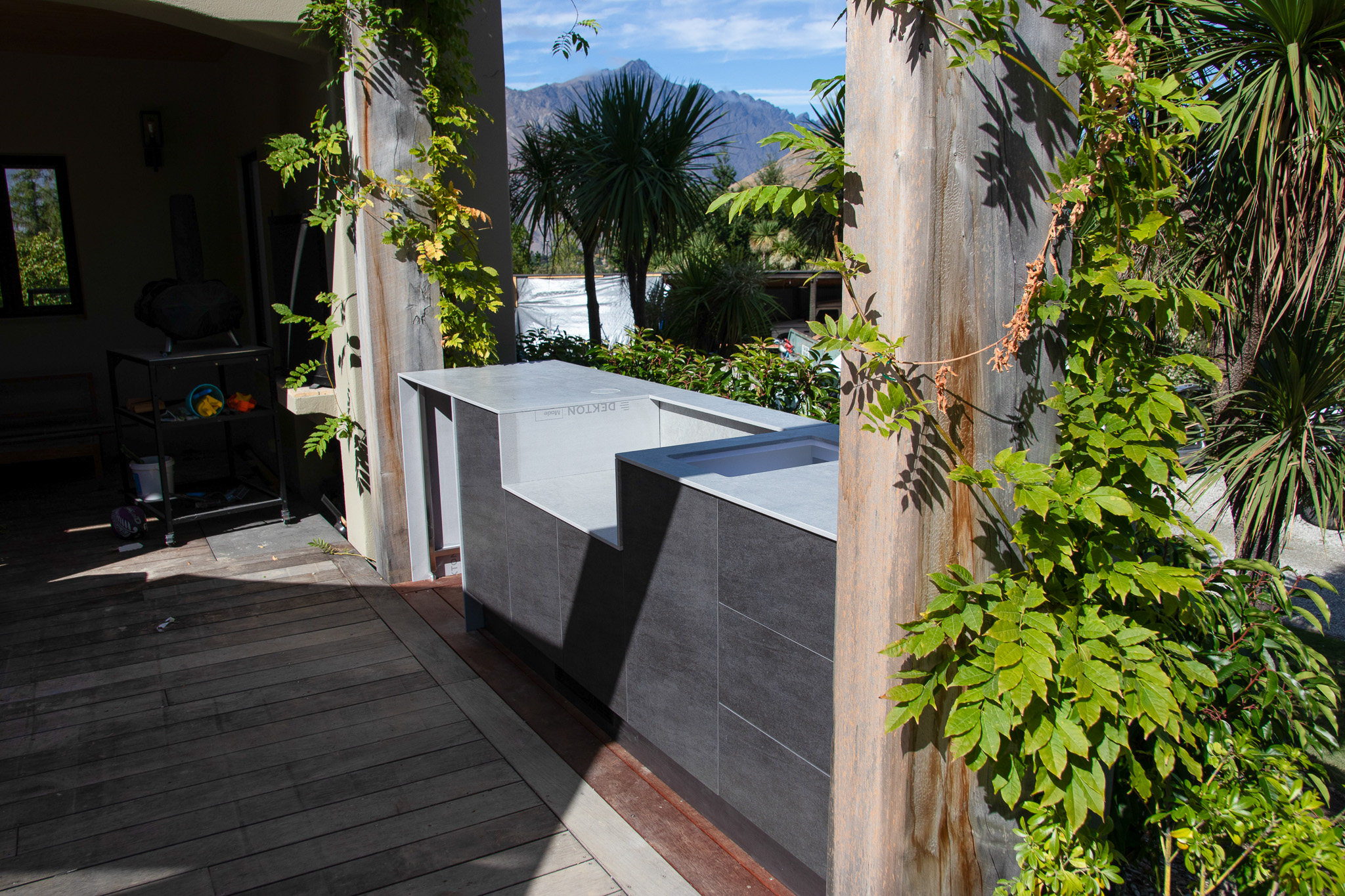 Modern outdoor kitchen with grey cabinets and a concrete countertop, surrounded by green plants and mountains in the background.