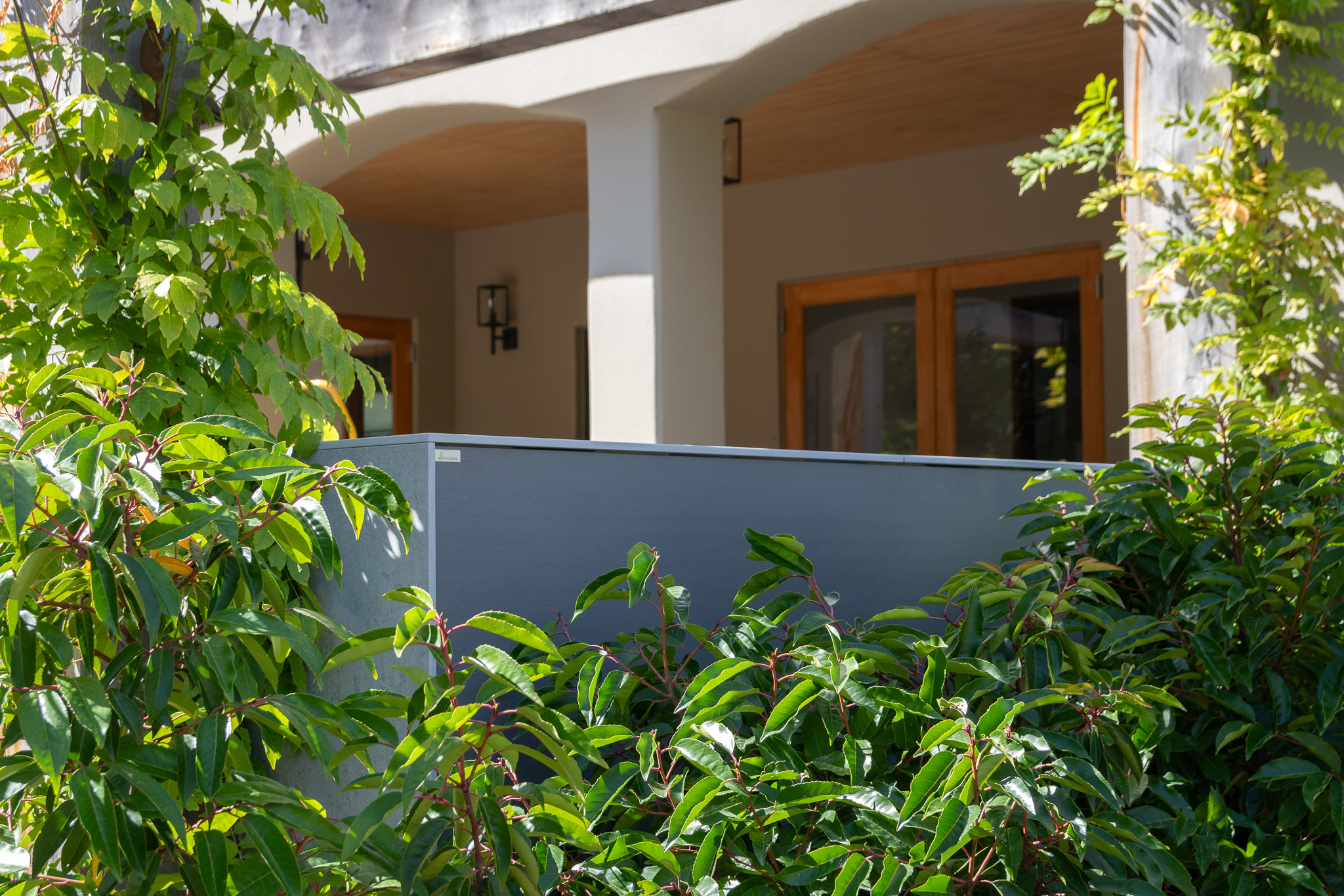 Modern outdoor patio with wooden framed glass doors behind lush green foliage and a sleek grey privacy screen.